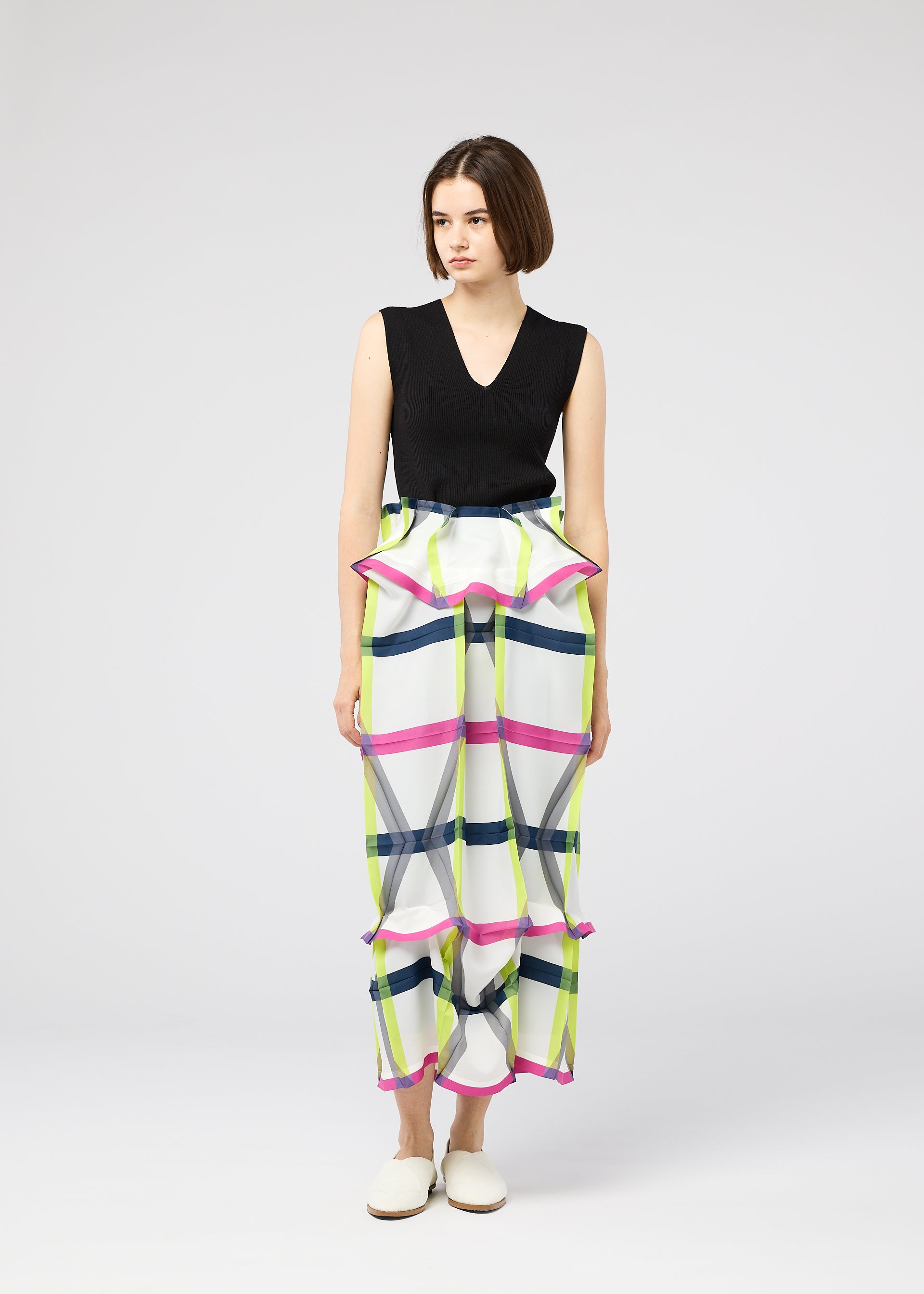 Zoomed view A woman with a bob haircut wears the NO.6 TRANSLUCENT by 132 5. ISSEY MIYAKE—sleeveless black top and bold plaid, high-waisted wide-leg pants with ruffles and folds—standing against a plain white background.