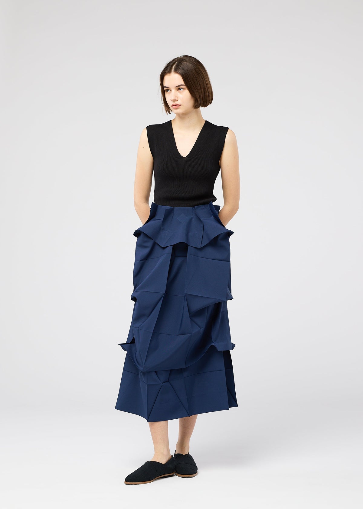 A woman with short brown hair stands against a plain background, wearing the NO.6 SOLID high-waisted blue skirt by 132 5. ISSEY MIYAKE, featuring geometric folds, paired with a sleeveless black top and black shoes, hands in her pockets. view 7 of 11