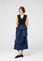 A woman with short brown hair stands against a plain background, wearing the NO.6 SOLID high-waisted blue skirt by 132 5. ISSEY MIYAKE, featuring geometric folds, paired with a sleeveless black top and black shoes, hands in her pockets.