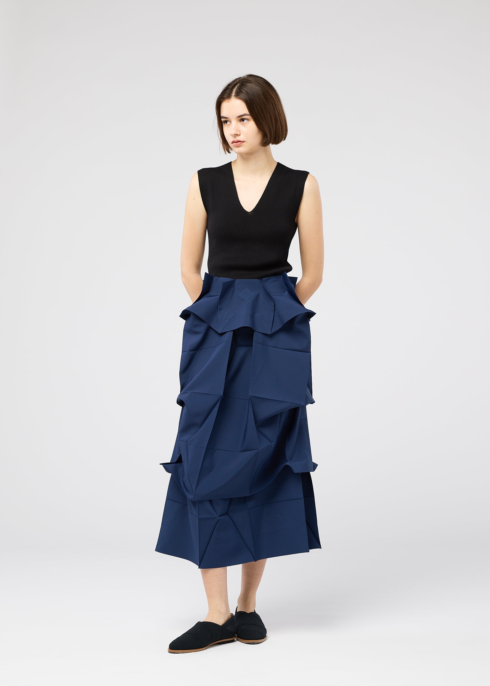 Zoomed view A woman with short brown hair stands against a plain background, wearing the NO.6 SOLID high-waisted blue skirt by 132 5. ISSEY MIYAKE, featuring geometric folds, paired with a sleeveless black top and black shoes, hands in her pockets.