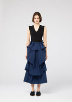 A woman with short brown hair wears the NO.6 SOLID skirt by 132 5. ISSEY MIYAKE, a blue ruffled tiered piece with a three-dimensional structure, paired with a sleeveless black top and black slip-on shoes, facing the camera neutrally.