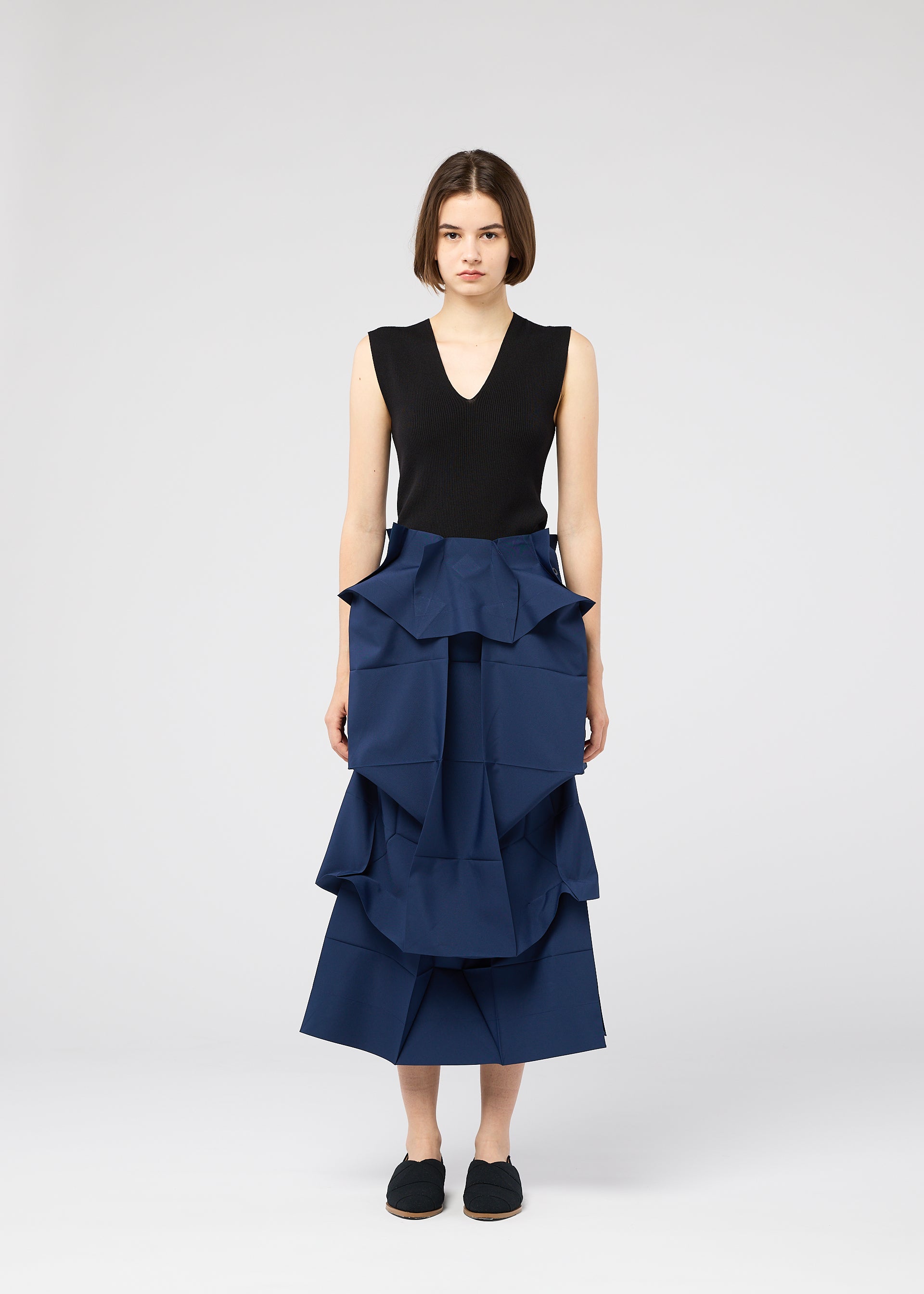 Zoomed view A woman with short brown hair wears the NO.6 SOLID skirt by 132 5. ISSEY MIYAKE, a blue ruffled tiered piece with a three-dimensional structure, paired with a sleeveless black top and black slip-on shoes, facing the camera neutrally.