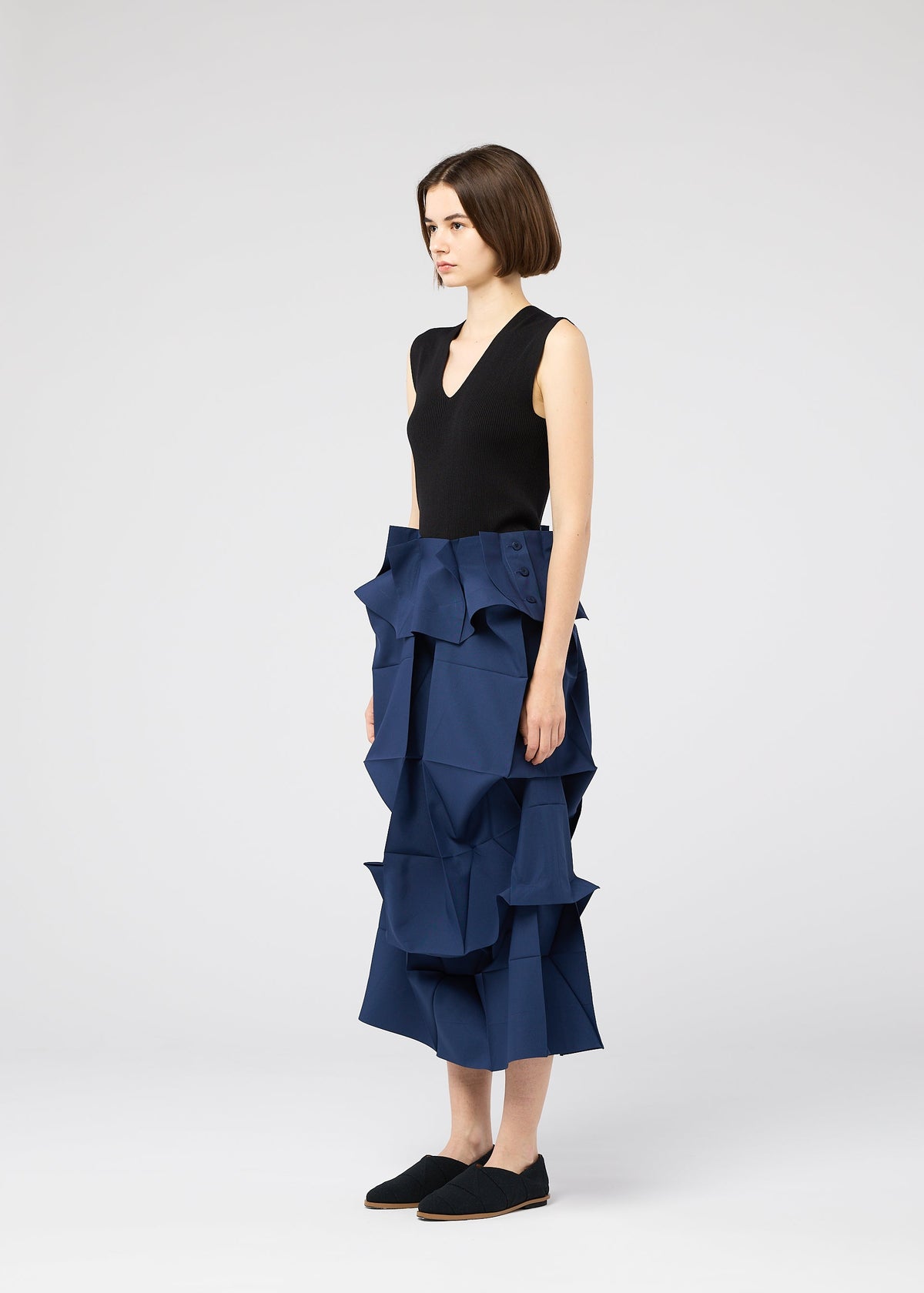 A woman in profile wears a sleeveless black top and the NO.6 SOLID skirt by 132 5. ISSEY MIYAKE, featuring bold blue geometric ruffles for a sculptural look, paired with black slip-on shoes against a plain background. view 9 of 11