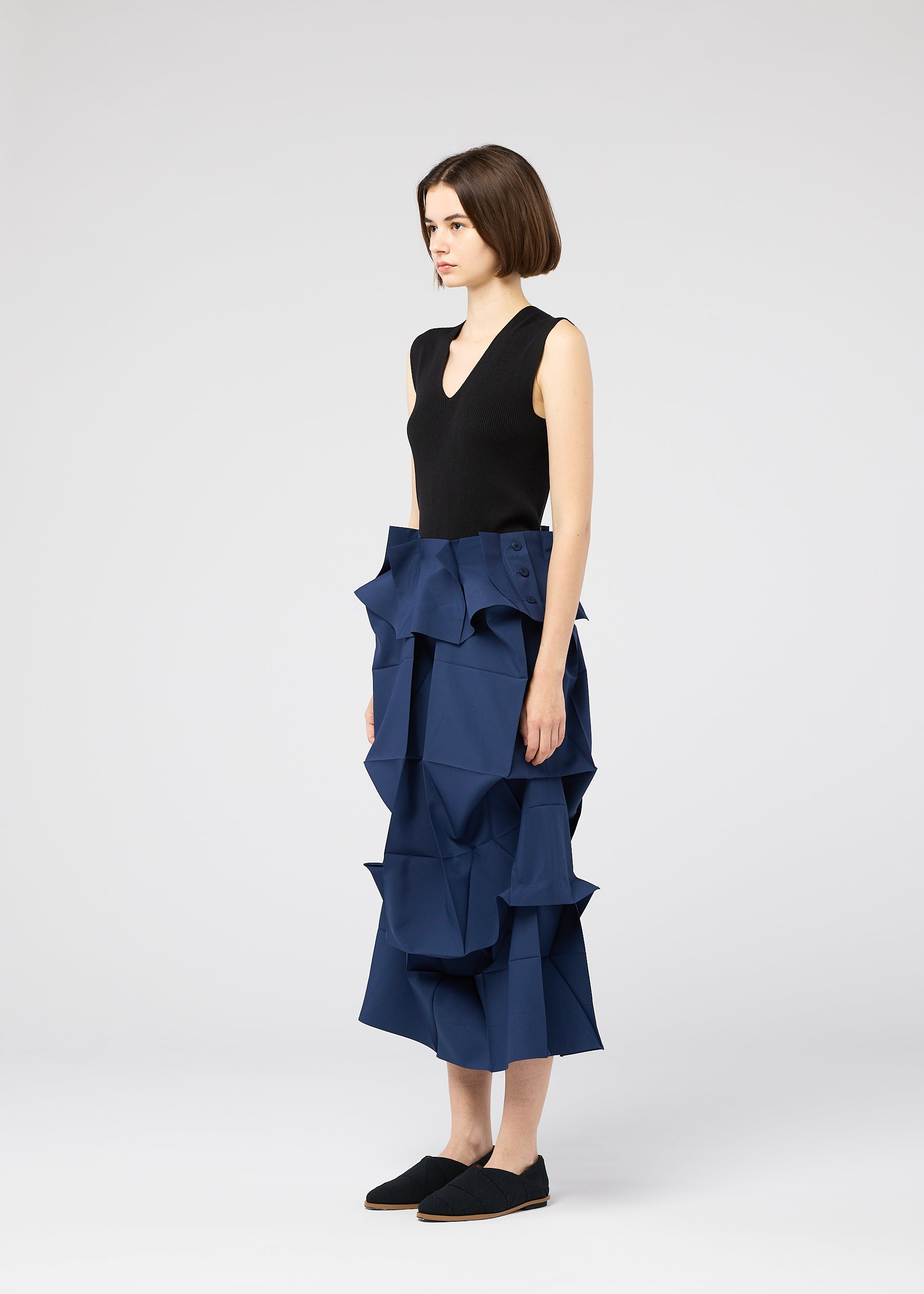Zoomed view A woman in profile wears a sleeveless black top and the NO.6 SOLID skirt by 132 5. ISSEY MIYAKE, featuring bold blue geometric ruffles for a sculptural look, paired with black slip-on shoes against a plain background.