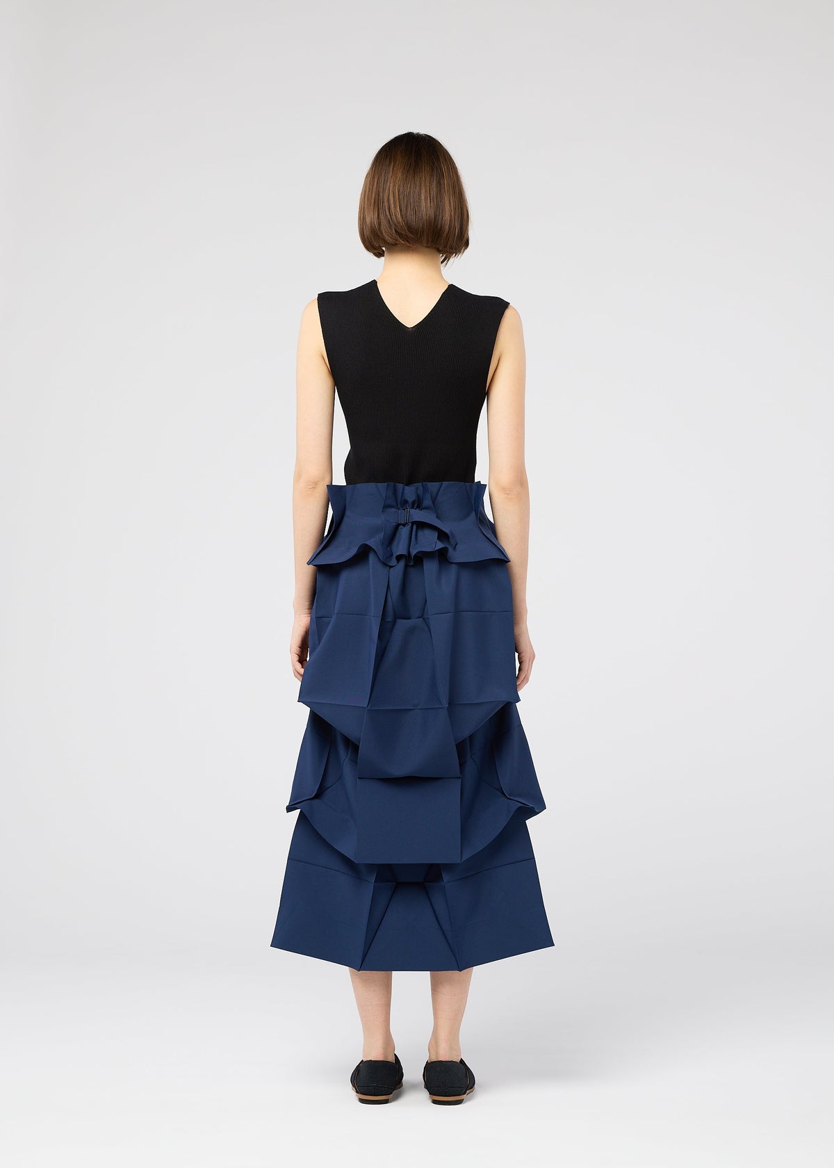 A person with short brown hair, seen from behind, wears a sleeveless black top and the 132 5. ISSEY MIYAKE NO.6 SOLID navy skirt with a waist bow, highlighting its three-dimensional shape against a plain light gray background. view 10 of 11