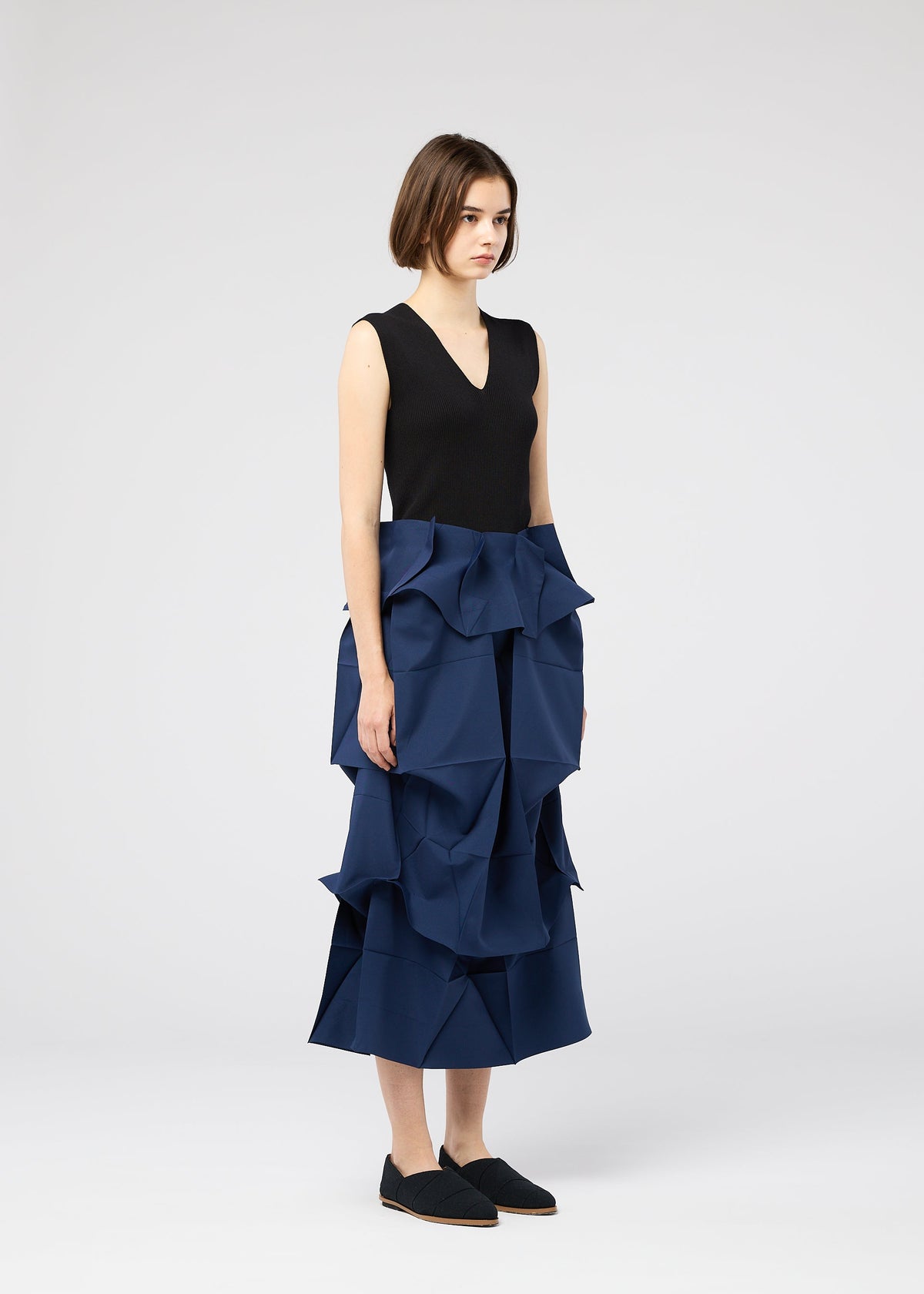 A woman with short brown hair wears the NO.6 SOLID blue midi skirt by 132 5. ISSEY MIYAKE, featuring large ruffles and a 3D structure, paired with a sleeveless black top and black slip-on shoes against a light gray background. view 11 of 11