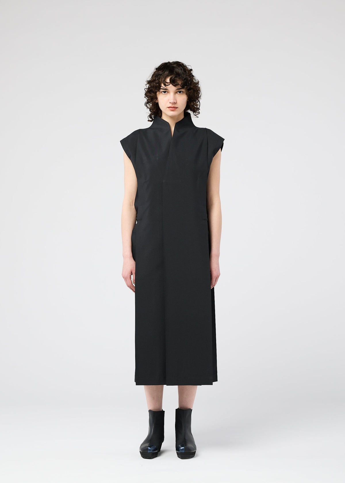 A person with short curly hair faces forward, wearing a sleeveless, ankle-length 132 5. FLAT 1 dress by 132 5. ISSEY MIYAKE and black ankle boots, against a plain light gray background. view 7 of 10