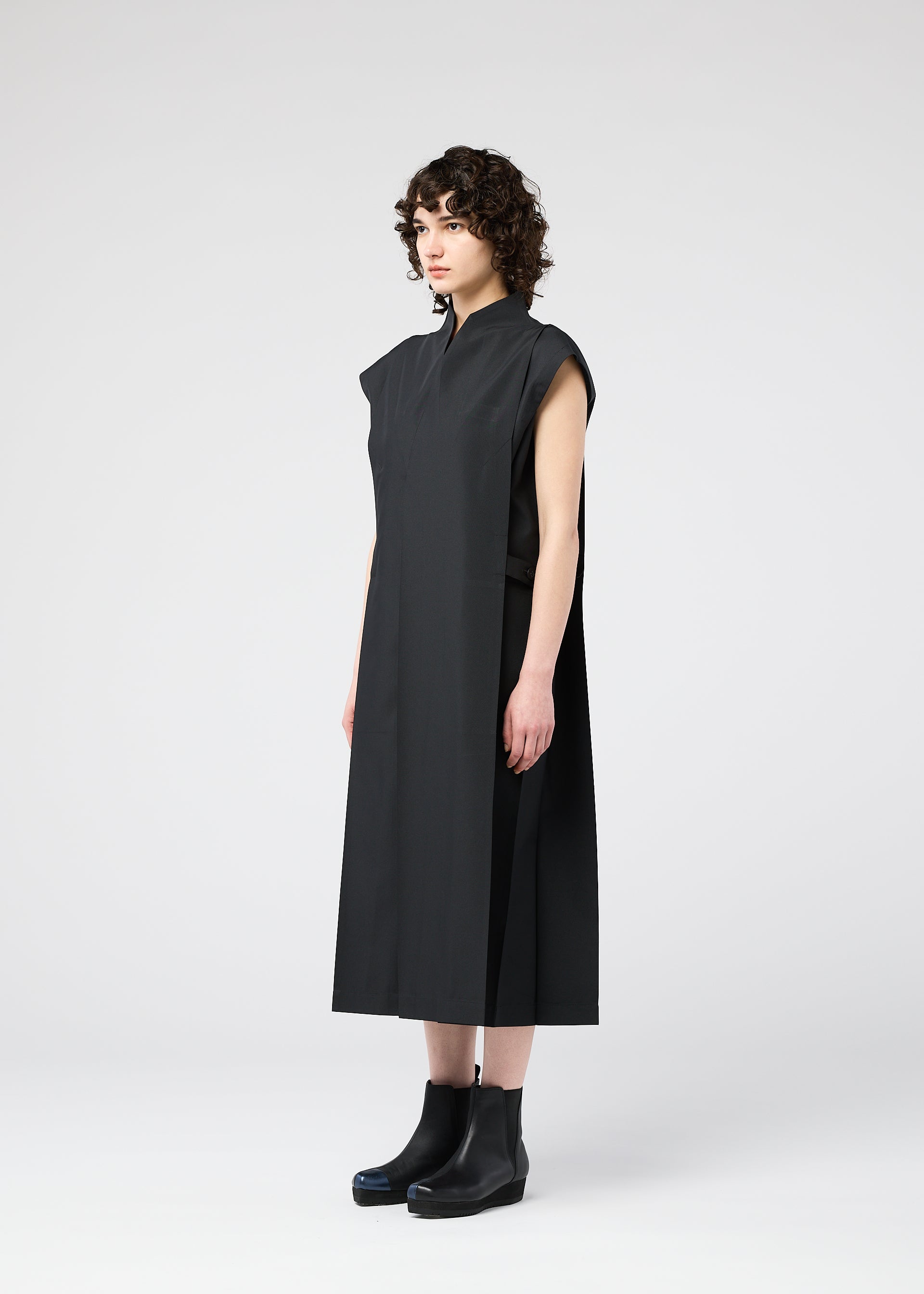 Zoomed view A person with short curly hair wears a sleeveless, loose-fit black midi dress—132 5. FLAT 1 by 132 5. ISSEY MIYAKE—and black ankle boots, standing against a plain light gray background.