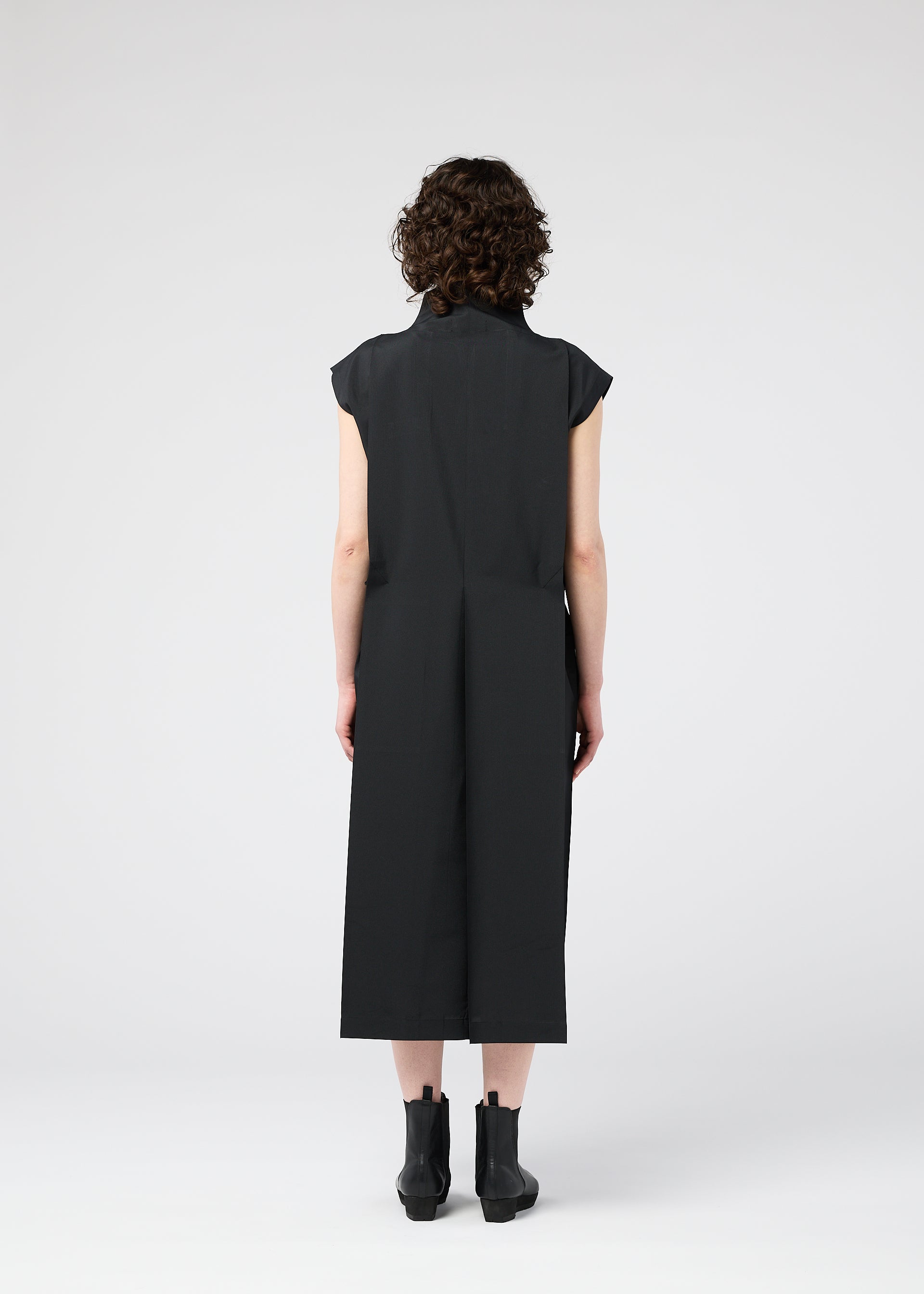 Zoomed view A person with short curly hair, wearing a black 132 5. FLAT 1 dress by 132 5. ISSEY MIYAKE—renowned for its unique garment structure—and black ankle boots stands facing away against a plain light background.