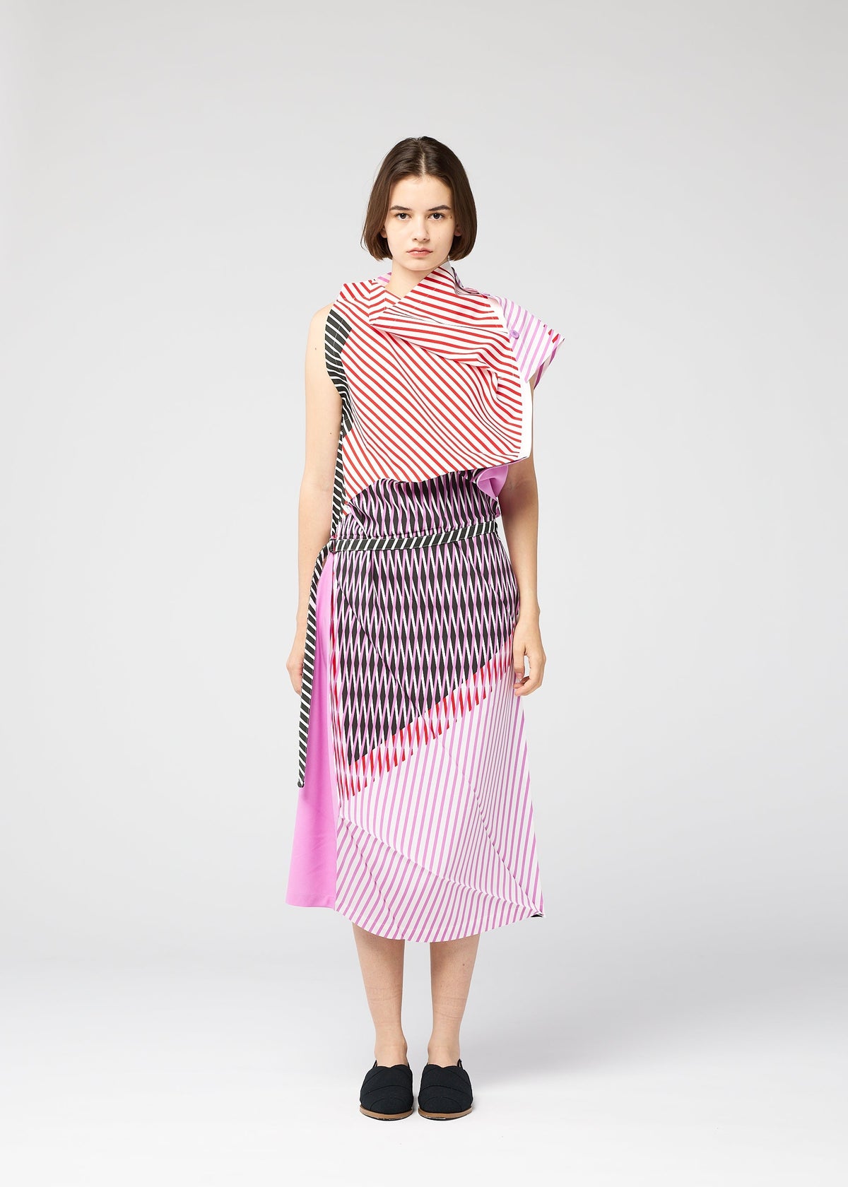 A person with short brown hair and black slip-on shoes stands against a plain background, wearing the FACET sleeveless dress by 132 5. ISSEY MIYAKE, featuring bold diagonal pink, red, black, and white stripes for a striking look. view 7 of 11