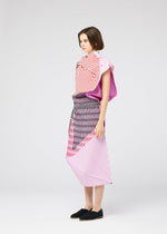 A woman with short brown hair and black slip-on shoes models the FACET set by 132 5. ISSEY MIYAKE: a bold pink, red, and white striped top and matching ankle-length geometric skirt, posing sideways against a plain background.