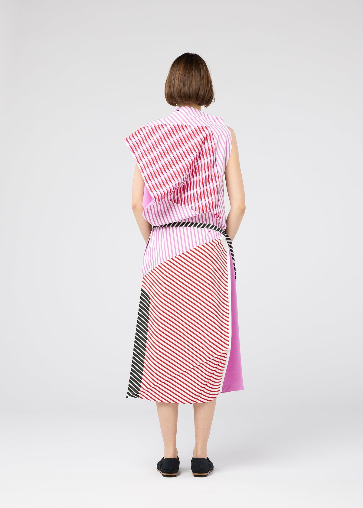 A woman faces away, wearing the FACET sleeveless calf-length dress by 132 5. ISSEY MIYAKE, featuring bold Nassen print stripes in red, pink, black, and white. She pairs it with black flats against a plain light gray background. view 9 of 11