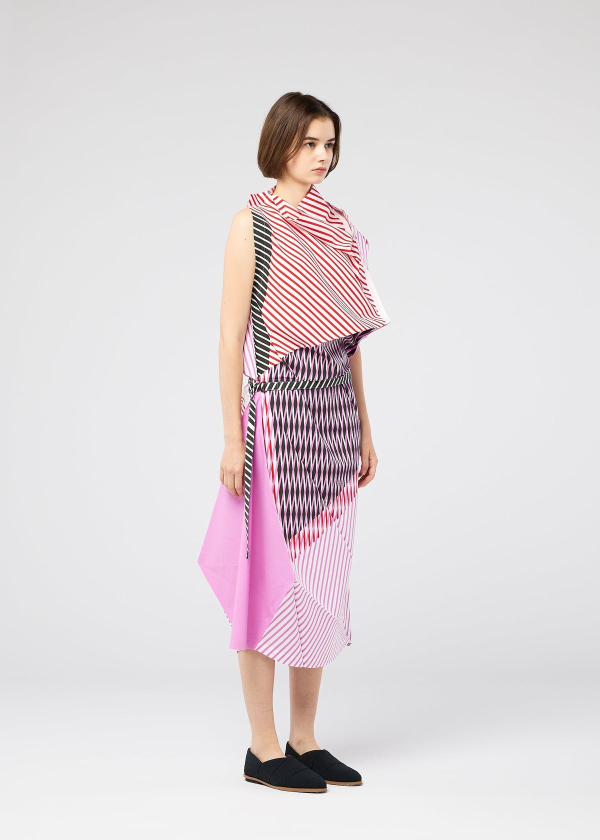 A woman wears FACET, a sleeveless asymmetrical dress by 132 5. ISSEY MIYAKE, featuring bold pink, black, and white geometric stripes in the Nassen print. She accessorizes with a black belt and slip-on shoes, her short brown hair completing the look. view 10 of 11