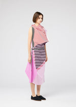 A woman wears FACET, a sleeveless asymmetrical dress by 132 5. ISSEY MIYAKE, featuring bold pink, black, and white geometric stripes in the Nassen print. She accessorizes with a black belt and slip-on shoes, her short brown hair completing the look.