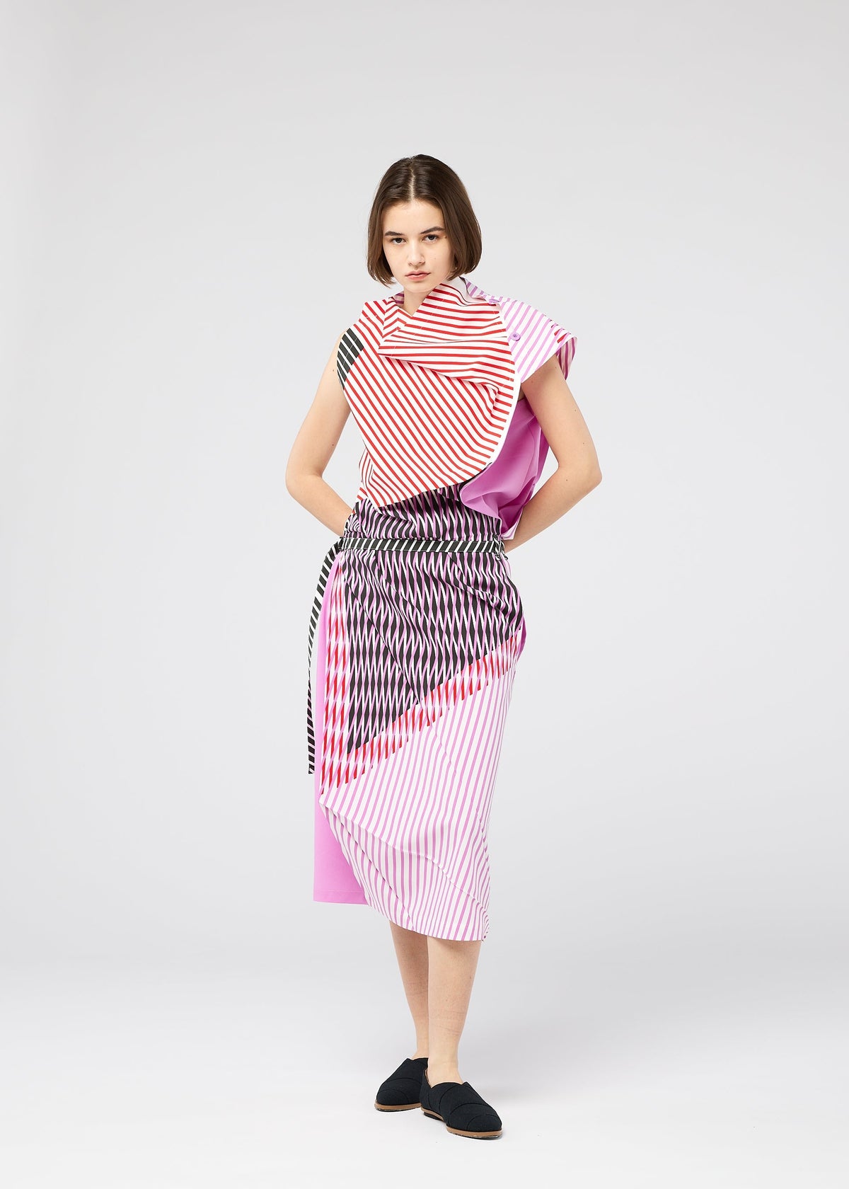 A person poses against a plain background, wearing FACET by 132 5. ISSEY MIYAKE—a colorful outfit with geometric, three-dimensional red, pink, black, and white striped Nassen print—paired with black shoes. view 11 of 11