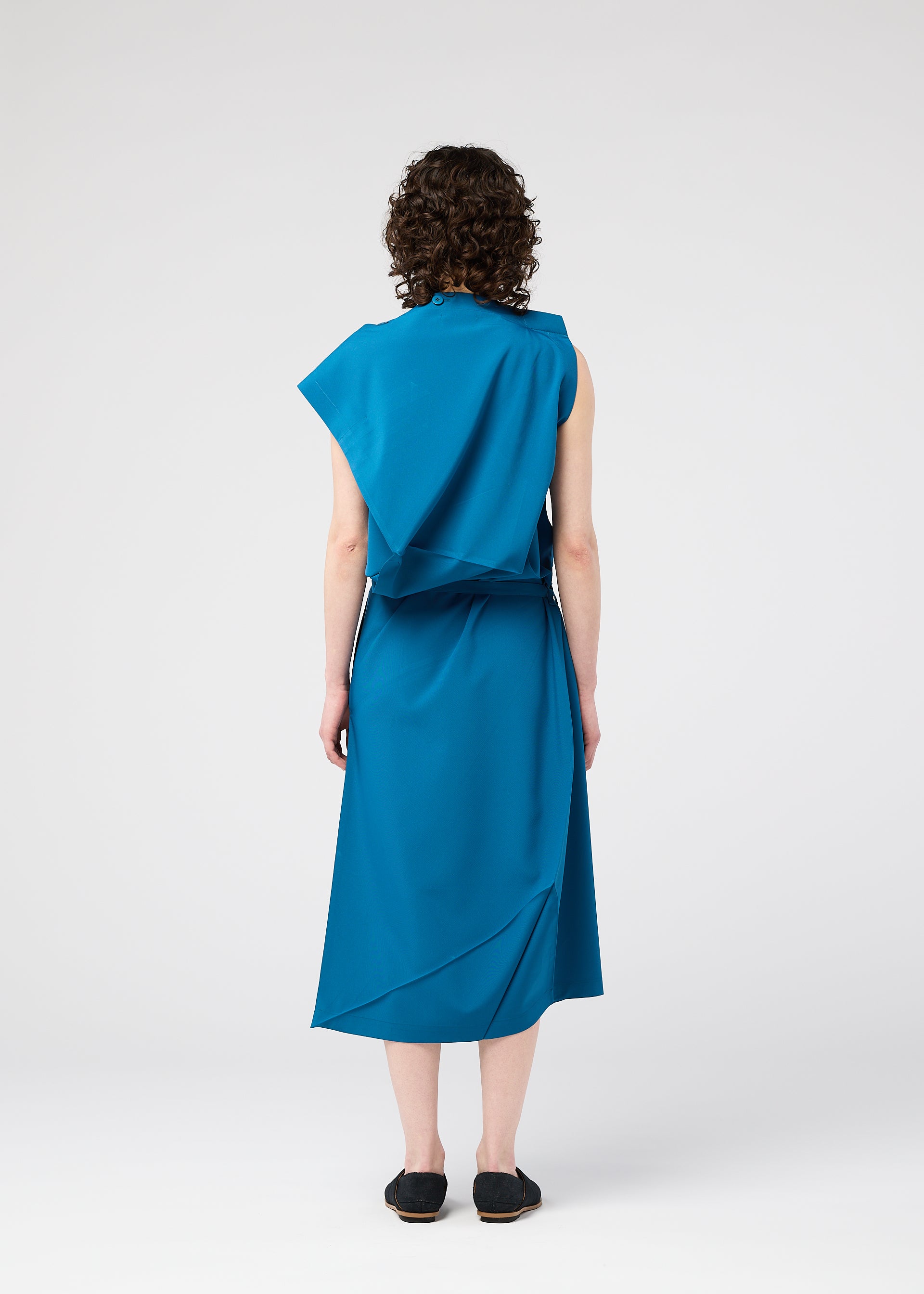Zoomed view A person with curly hair stands facing away, wearing the FACET SOLID sleeveless teal asymmetrical midi dress by 132 5. ISSEY MIYAKE, crafted from plant-derived polyester plain fabric, and black flat shoes against a plain light background.
