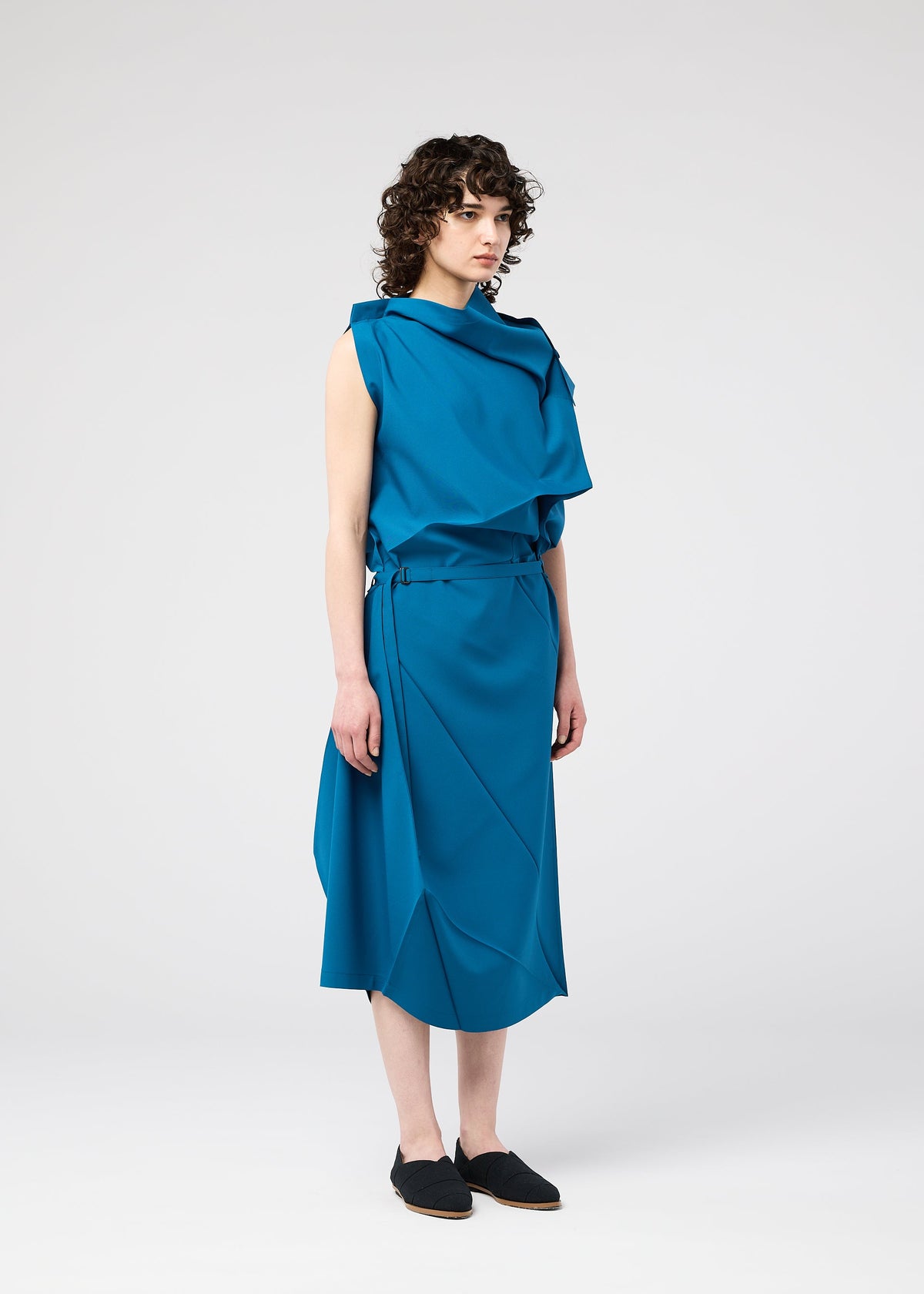 A person with curly brown hair stands in a studio wearing a loose, sleeveless blue FACET SOLID dress by 132 5. ISSEY MIYAKE, made from plant-derived polyester with draped details and black slip-on shoes against a plain white background. view 10 of 11