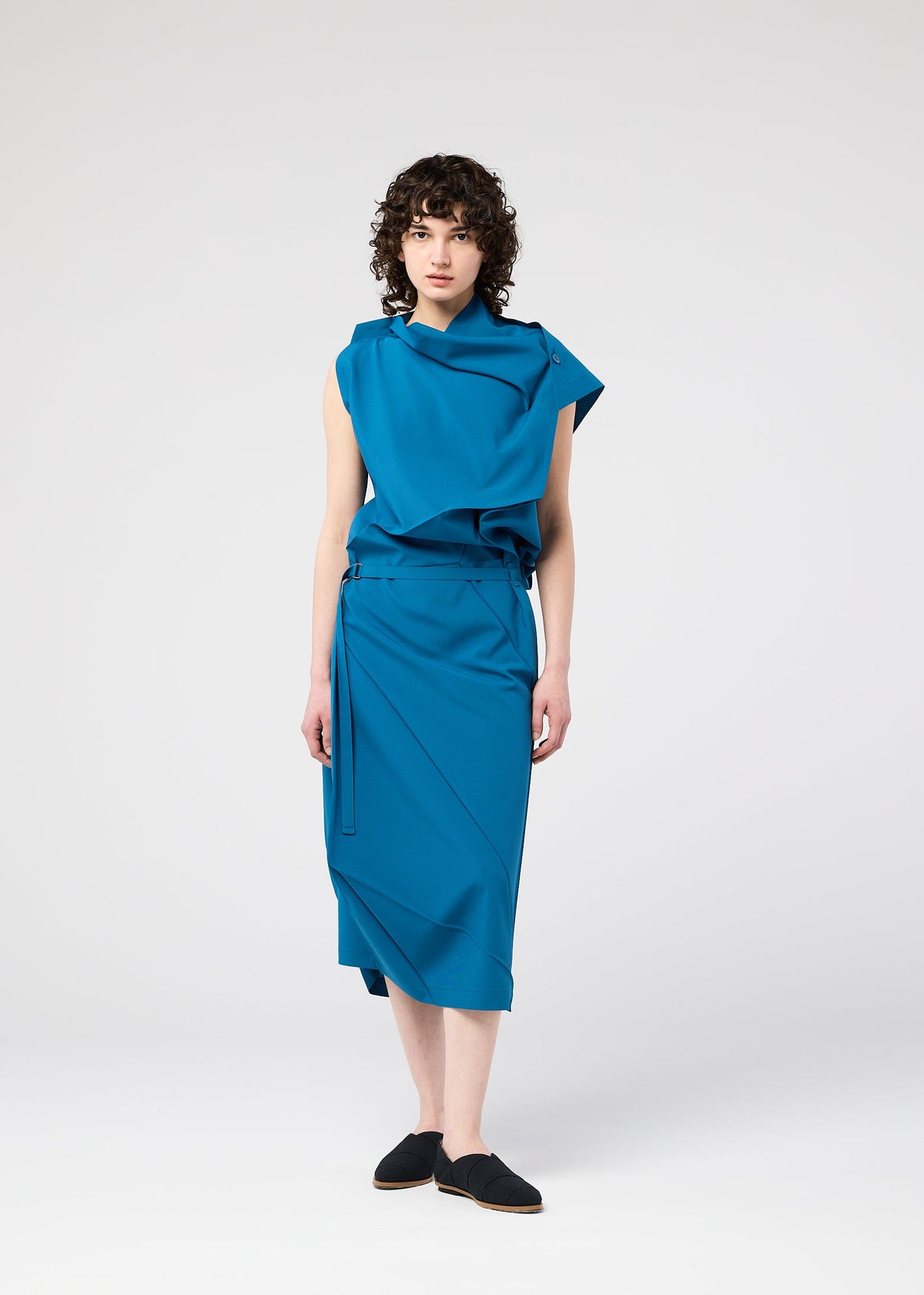 A person with curly brown hair wears the FACET SOLID sleeveless draped teal dress by 132 5. ISSEY MIYAKE, featuring an asymmetrical neckline and plant-derived polyester, paired with black slip-on shoes against a plain white background. view 11 of 11