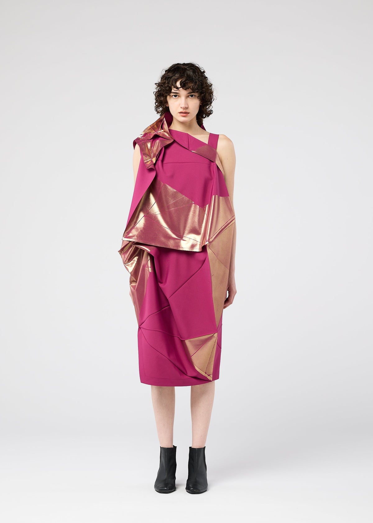 A woman with short curly hair wears the NO.17 FOIL sleeveless magenta dress by 132 5. ISSEY MIYAKE, featuring a shiny metallic gold foil print. She stands facing forward against a plain light background with black ankle boots. view 7 of 13