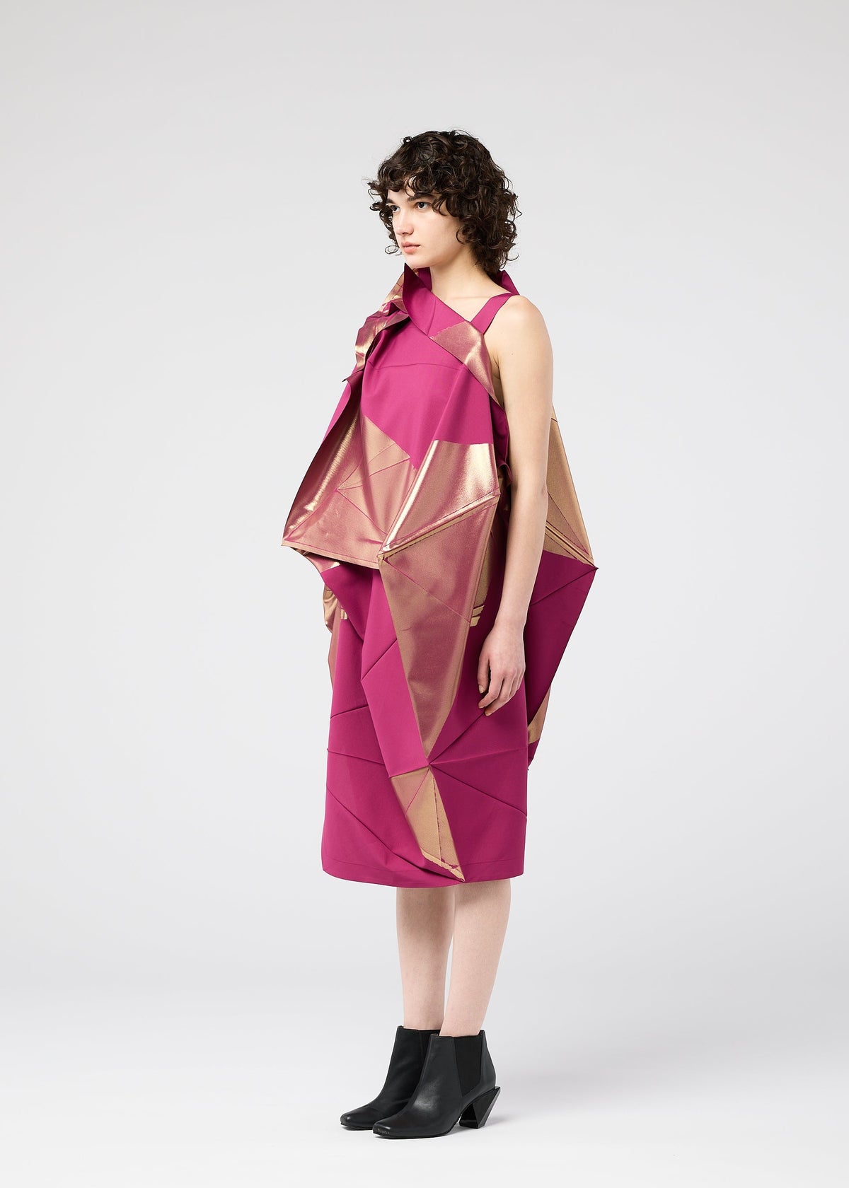 A woman in profile wears the NO.17 FOIL dress by 132 5. ISSEY MIYAKE, showcasing innovative magenta and gold angular folds, paired with black ankle boots, against a plain white background. view 8 of 13