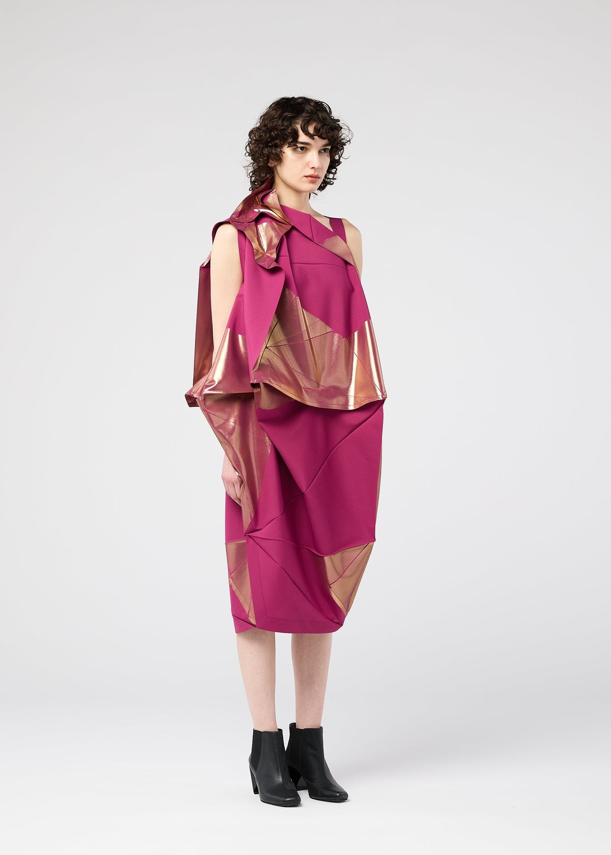 A person with short curly hair wears the 132 5. ISSEY MIYAKE NO.17 FOIL folding dress in bright magenta with geometric gold details and black ankle boots, creating a striking three-dimensional look against a plain light gray background. view 10 of 13