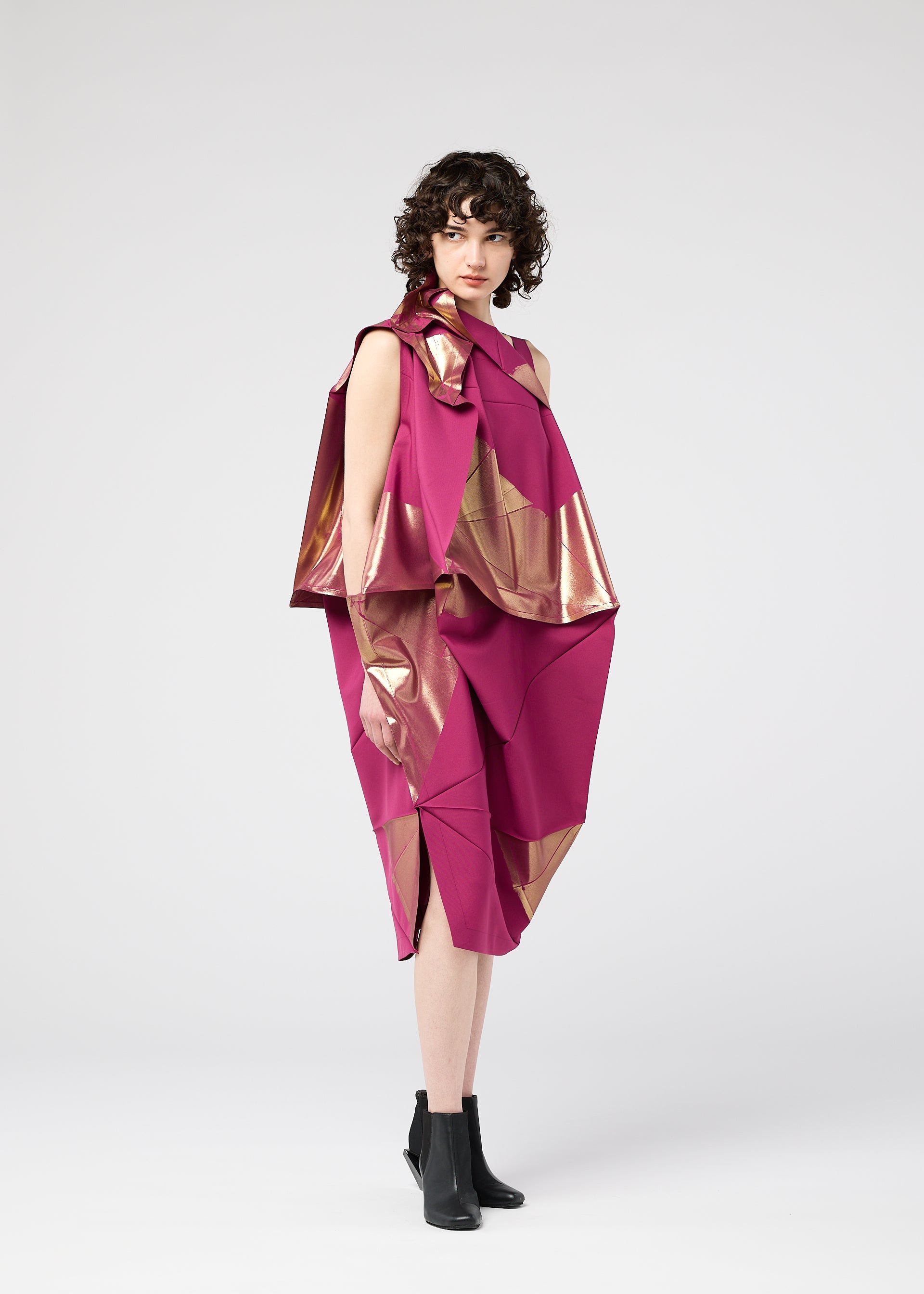 Zoomed view A woman with curly hair wears the 132 5. ISSEY MIYAKE NO.17 FOIL folding dress in magenta with metallic gold accents and black ankle boots, showcasing a three-dimensional silhouette against a plain light gray background.