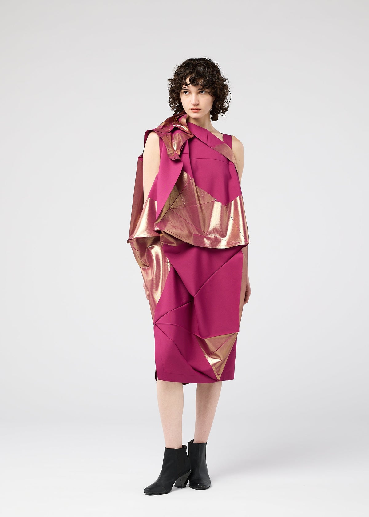 A woman with short curly hair wears the NO.17 FOIL folding dress by 132 5. ISSEY MIYAKE in magenta, featuring geometric gold foil accents and paired with black ankle boots, standing against a plain light gray background. view 12 of 13