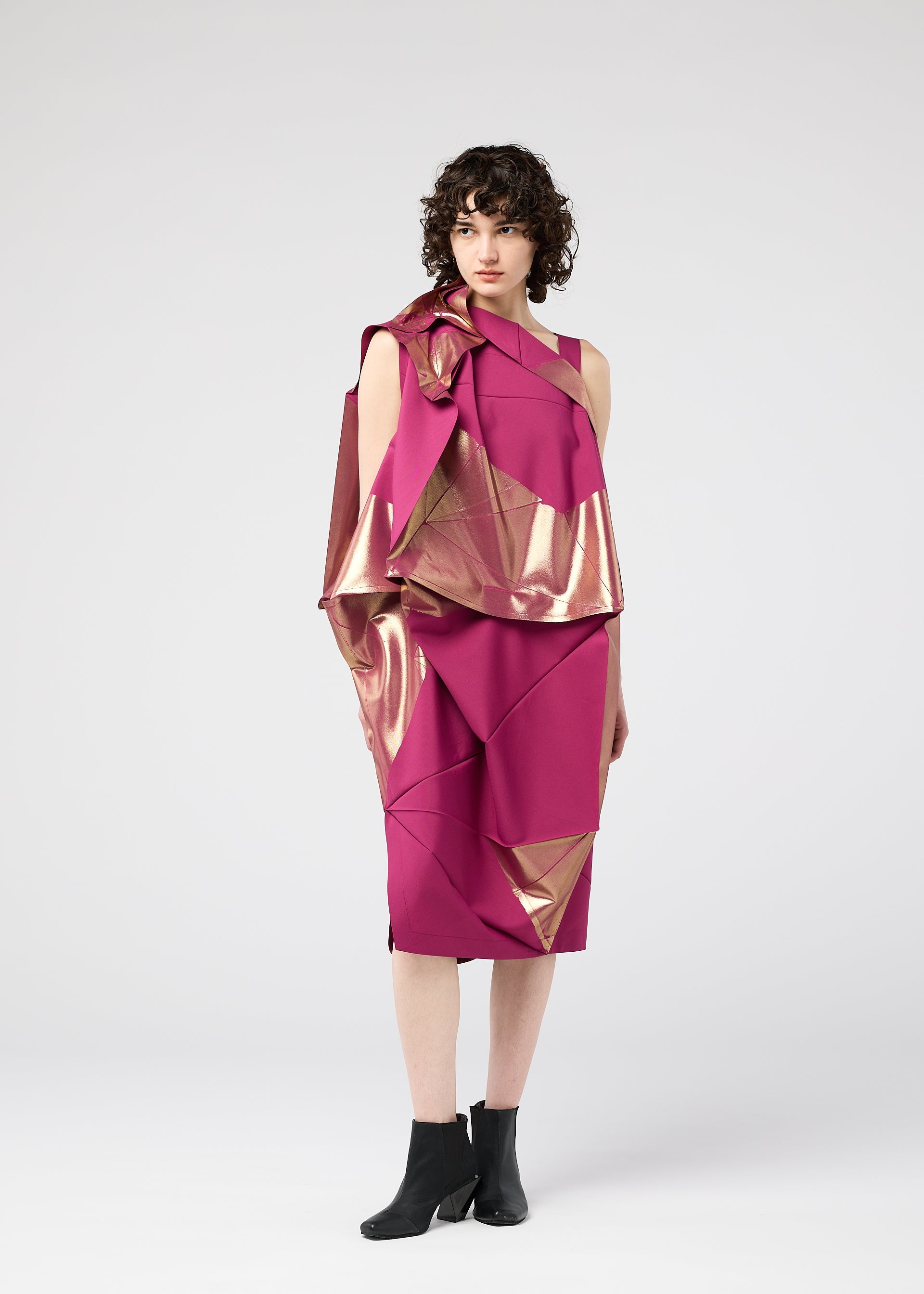 Zoomed view A woman with short curly hair wears the NO.17 FOIL folding dress by 132 5. ISSEY MIYAKE in magenta, featuring geometric gold foil accents and paired with black ankle boots, standing against a plain light gray background.