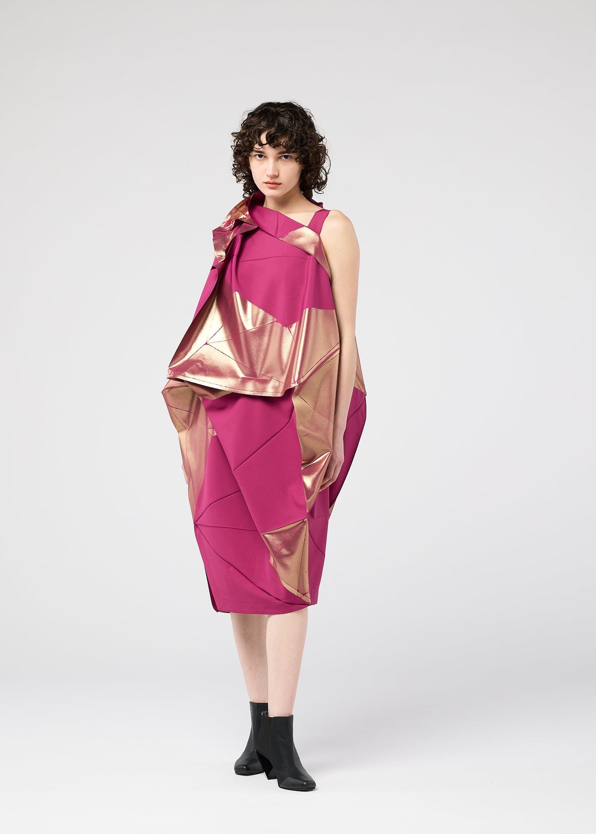 A woman with short curly hair models the 132 5. ISSEY MIYAKE NO.17 FOIL dress—an innovative pink and metallic gold geometric design with a three-dimensional silhouette—paired with black ankle boots against a light gray background. view 13 of 13