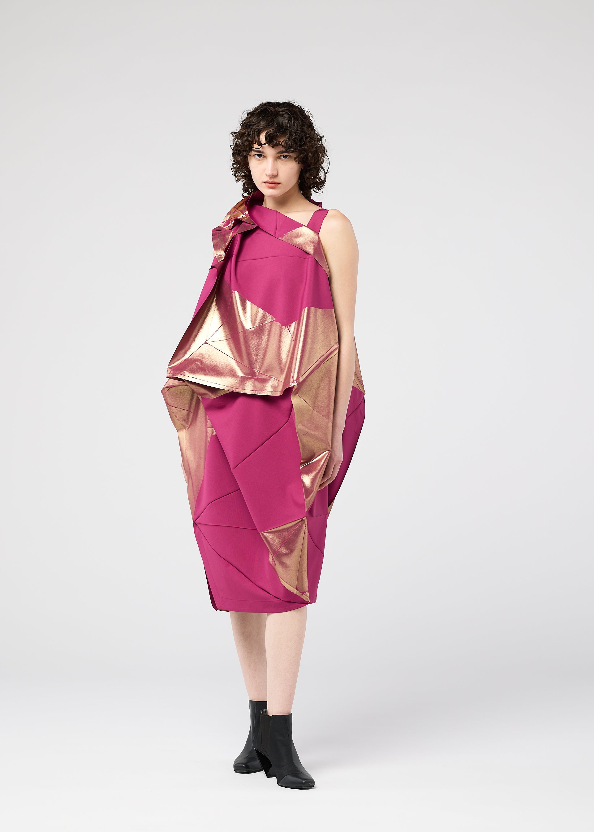 Zoomed view A woman with short curly hair models the 132 5. ISSEY MIYAKE NO.17 FOIL dress—an innovative pink and metallic gold geometric design with a three-dimensional silhouette—paired with black ankle boots against a light gray background.