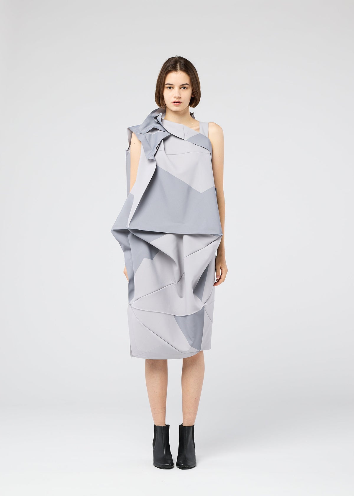 A woman faces forward in a geometric light grey NO.17 PRINT dress by 132 5. ISSEY MIYAKE, featuring angular folds and layers, paired with black ankle boots against a plain white background. view 7 of 11