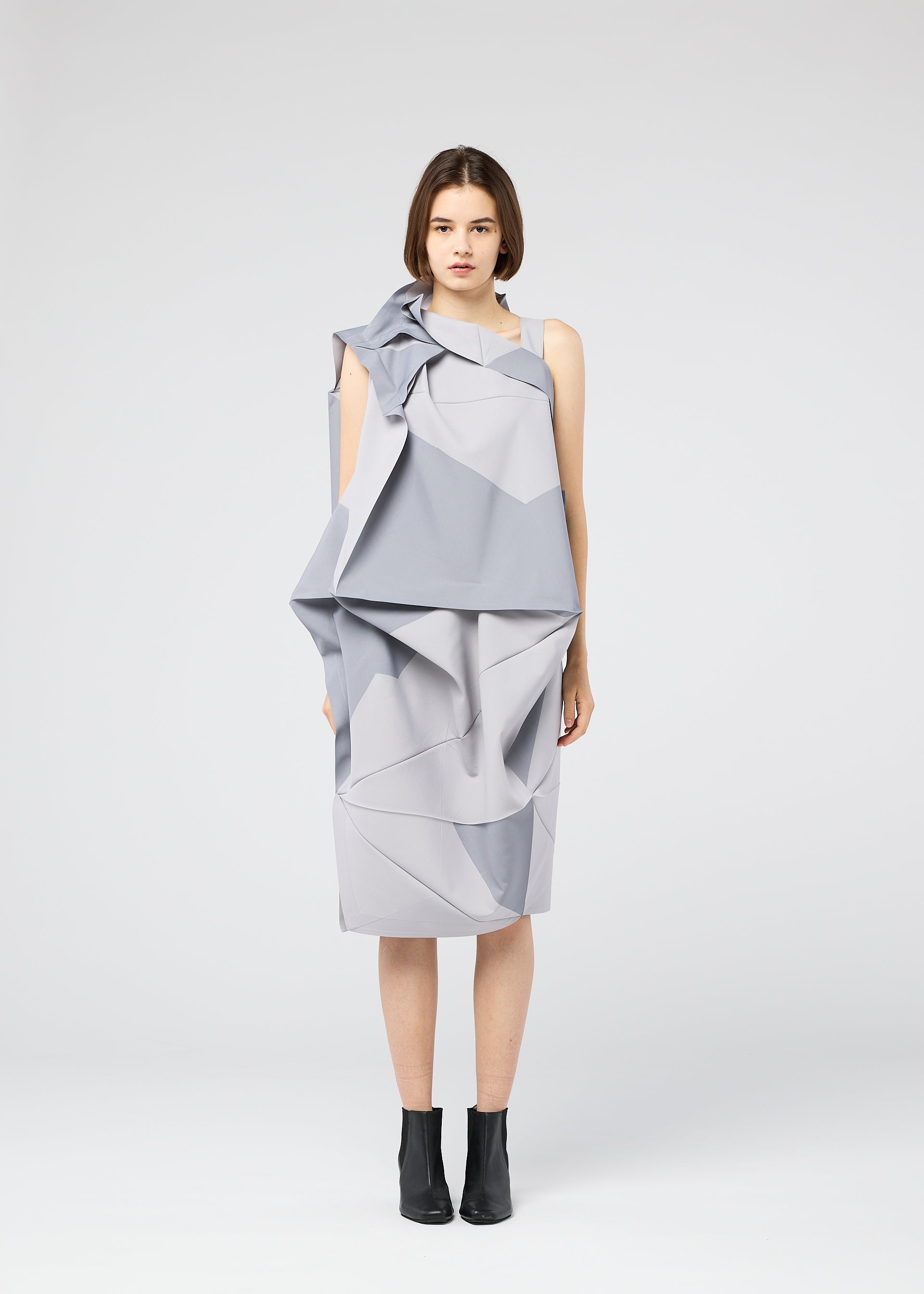 Zoomed view A woman faces forward in a geometric light grey NO.17 PRINT dress by 132 5. ISSEY MIYAKE, featuring angular folds and layers, paired with black ankle boots against a plain white background.