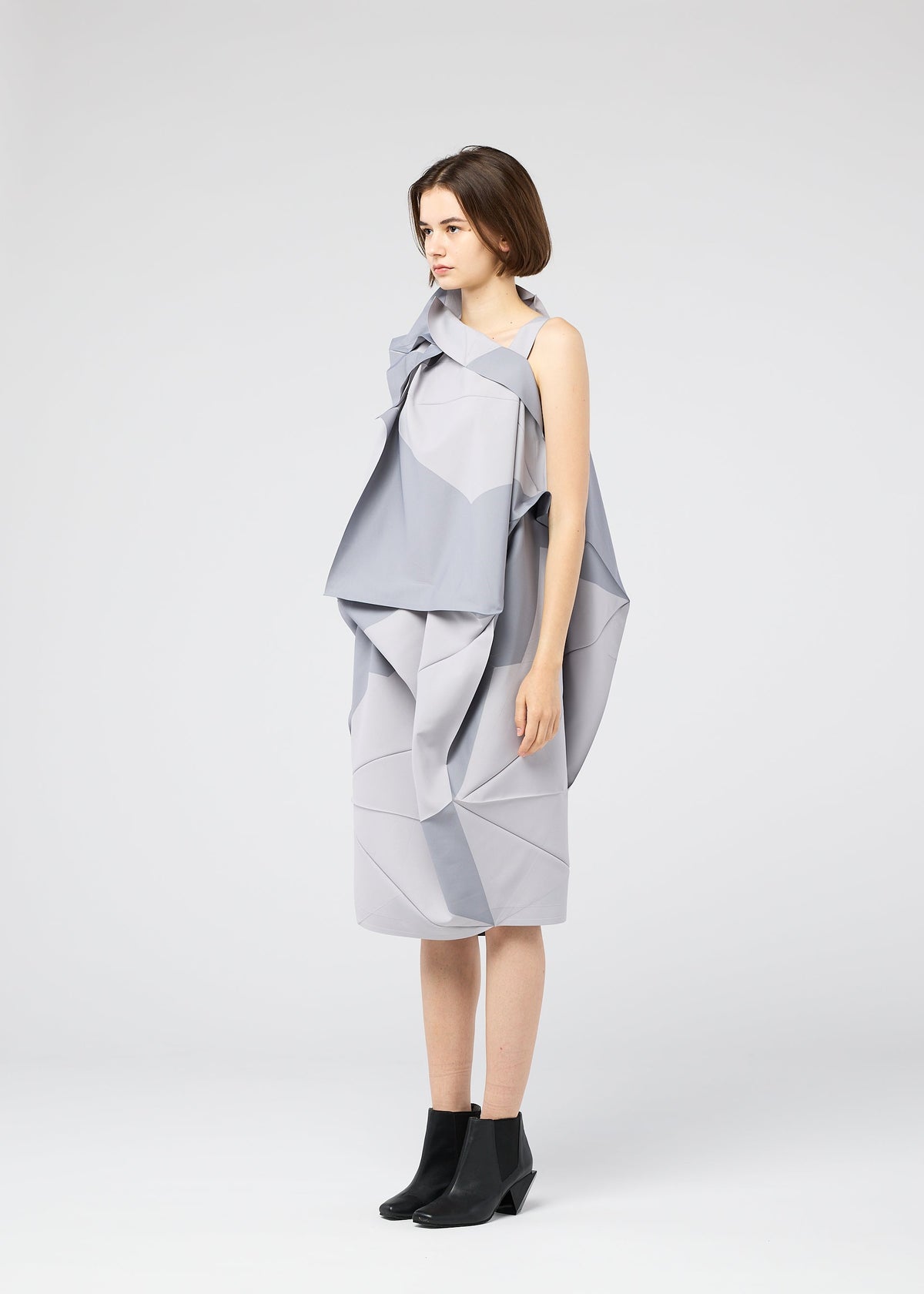 A woman stands in profile against a white background, wearing the NO.17 PRINT dress by 132 5. ISSEY MIYAKE—featuring sculptural, layered folds in light gray—and black ankle boots. view 8 of 11