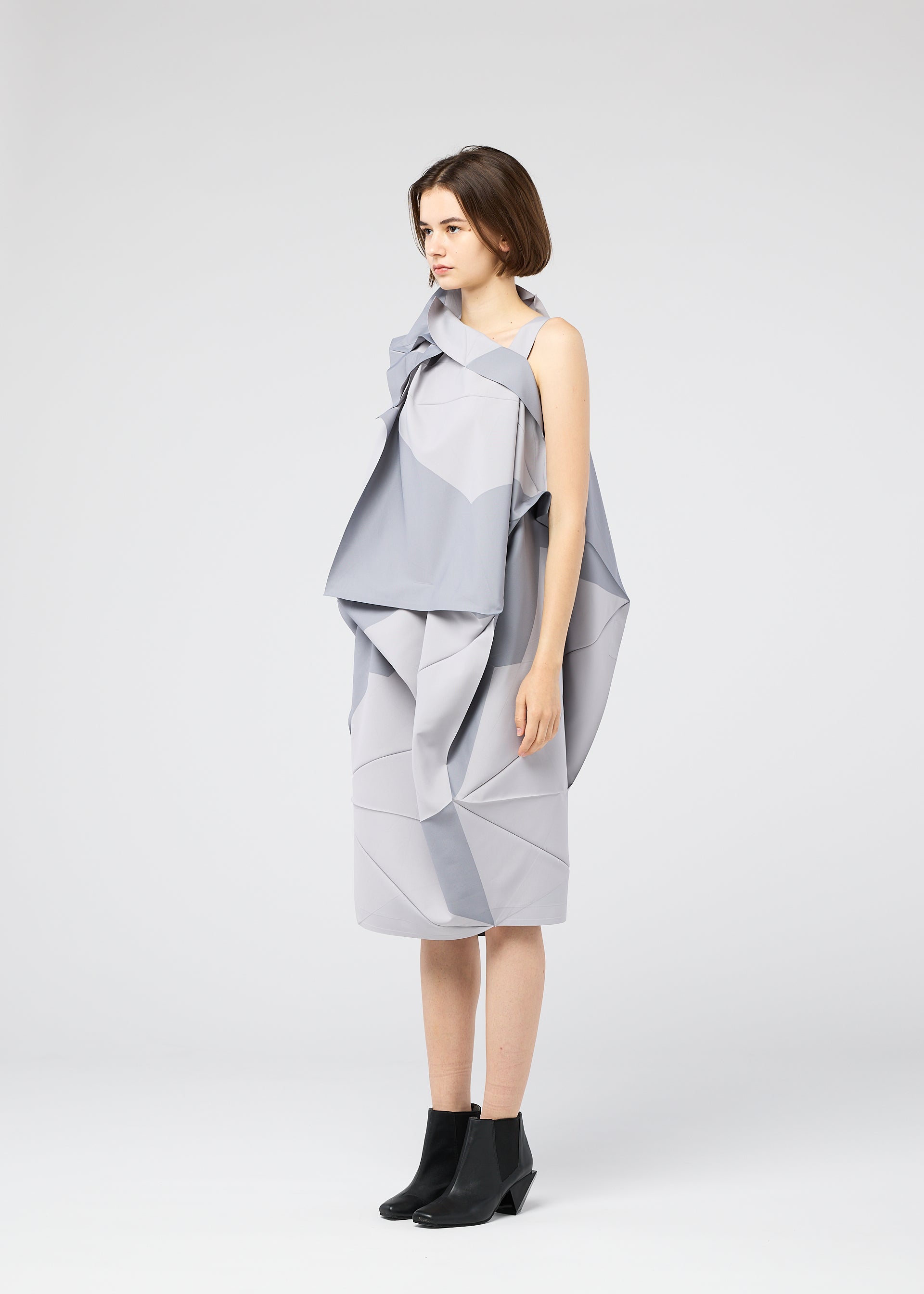 Zoomed view A woman stands in profile against a white background, wearing the NO.17 PRINT dress by 132 5. ISSEY MIYAKE—featuring sculptural, layered folds in light gray—and black ankle boots.