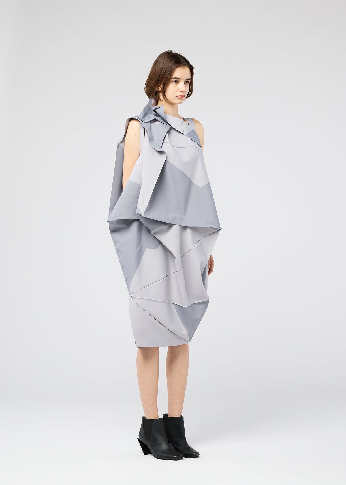 A woman with short brown hair stands against a plain light background, wearing the NO.17 PRINT dress by 132 5. ISSEY MIYAKE in light gray with angular folds and layers, paired with black ankle boots. view 10 of 11