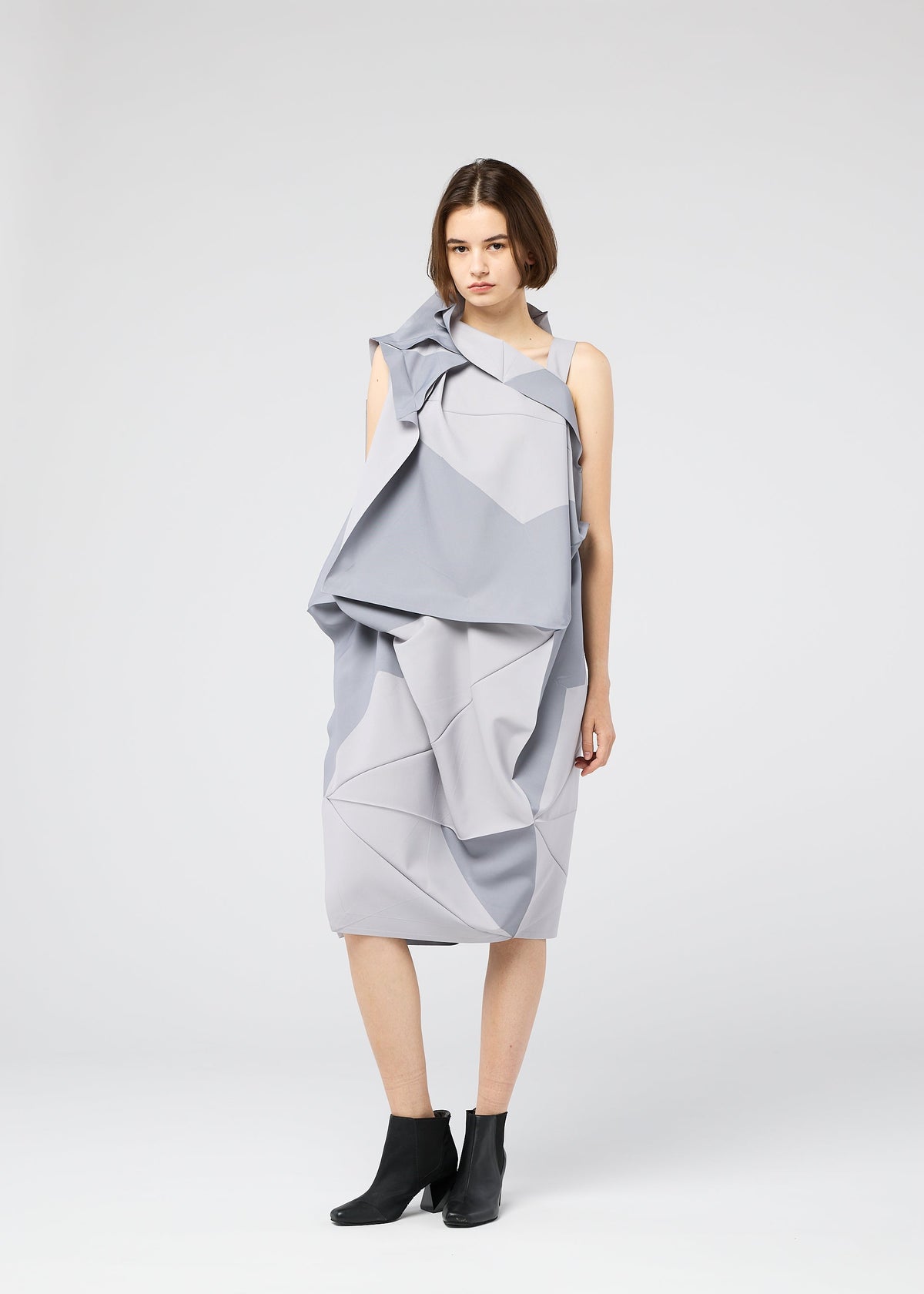 A woman stands against a plain background wearing the NO.17 PRINT dress by 132 5. ISSEY MIYAKE, featuring geometric folds and draping in light gray, paired with black ankle boots. She faces the camera neutrally, arms relaxed at her sides. view 11 of 11