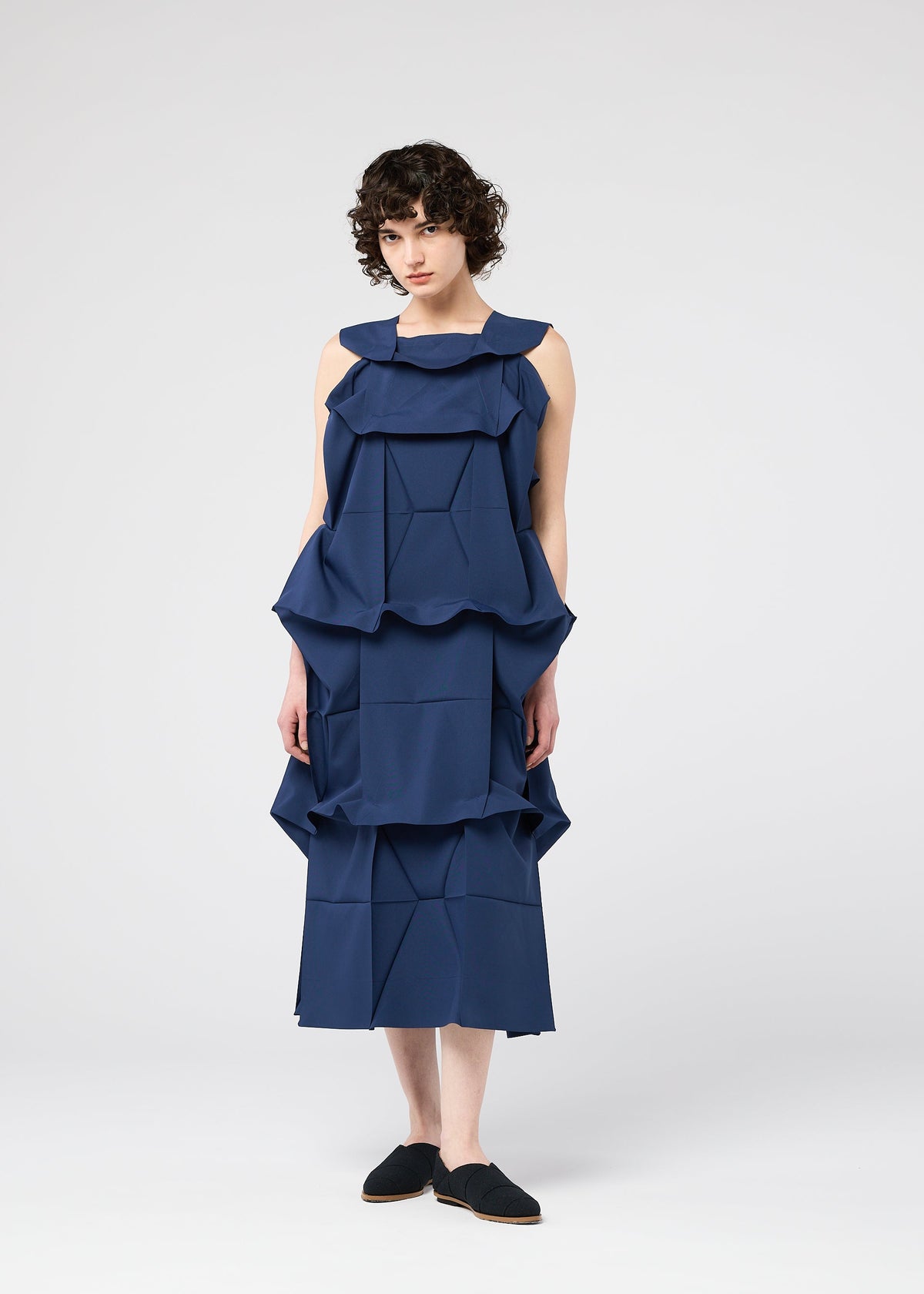 A person with short curly hair wears the sleeveless navy NO.6 SOLID dress by 132 5. ISSEY MIYAKE, featuring 3D geometric ruffles below the knees. They face forward against a white background, wearing black slip-on shoes. view 5 of 9