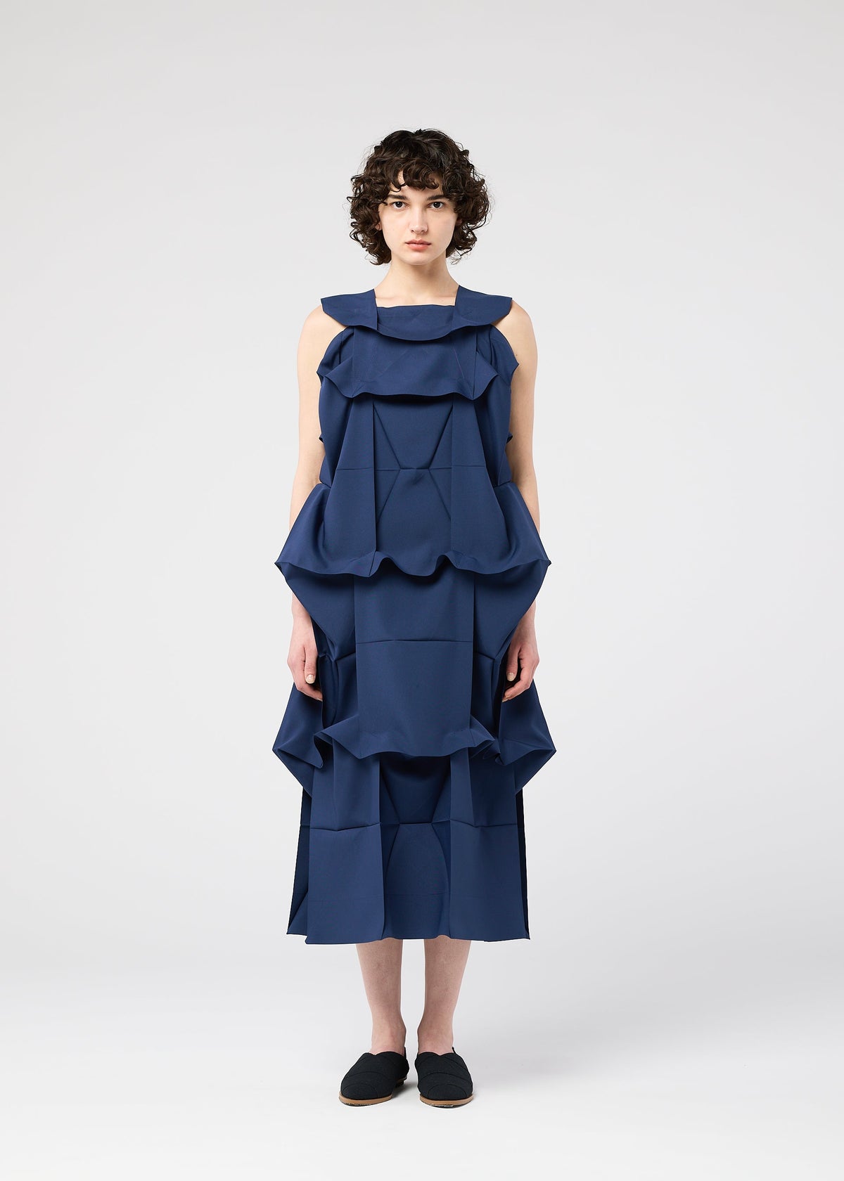 A person with short curly hair stands against a plain white background, wearing the NO.6 SOLID dress by 132 5. ISSEY MIYAKE, featuring layered sculptural ruffles for a striking three-dimensional look, paired with black slip-on shoes. view 6 of 9
