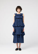 A person with short curly hair stands against a plain white background, wearing the NO.6 SOLID dress by 132 5. ISSEY MIYAKE, featuring layered sculptural ruffles for a striking three-dimensional look, paired with black slip-on shoes.