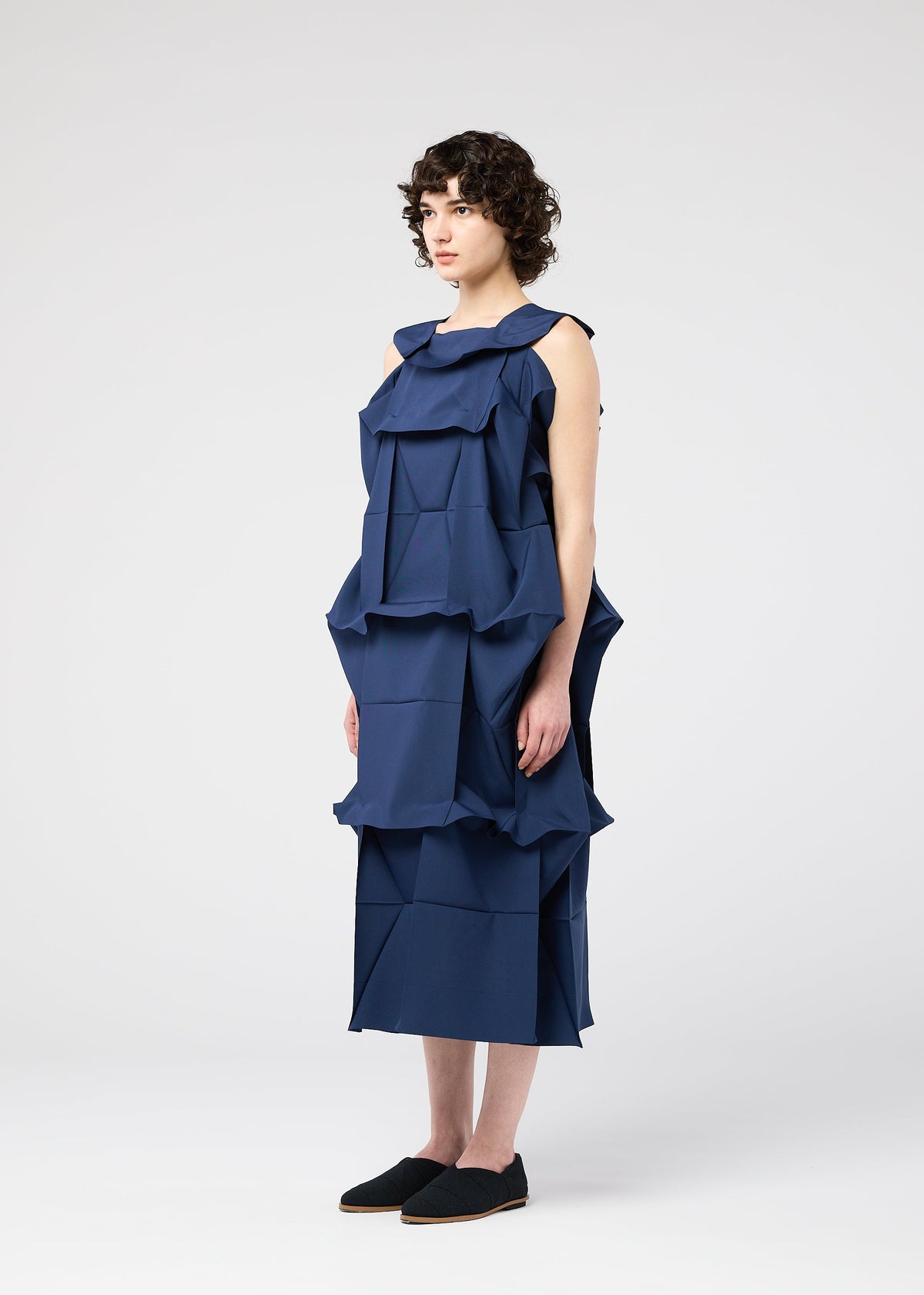 A woman with short, curly hair stands in a studio, wearing the sleeveless NO.6 SOLID dress by 132 5. ISSEY MIYAKE. The layered navy geometric panels create a three-dimensional look. She faces sideways in black slip-ons against a plain backdrop. view 7 of 9