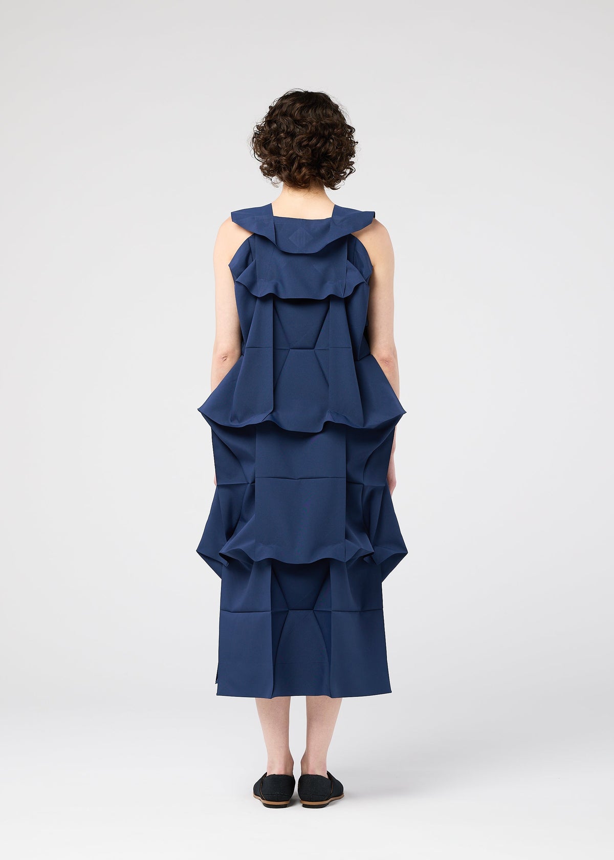 A person with short curly hair faces away wearing the 132 5. ISSEY MIYAKE NO.6 SOLID dress, which features large, structured geometric ruffles and a three-dimensional silhouette, with black slip-on shoes against a white background. view 8 of 9