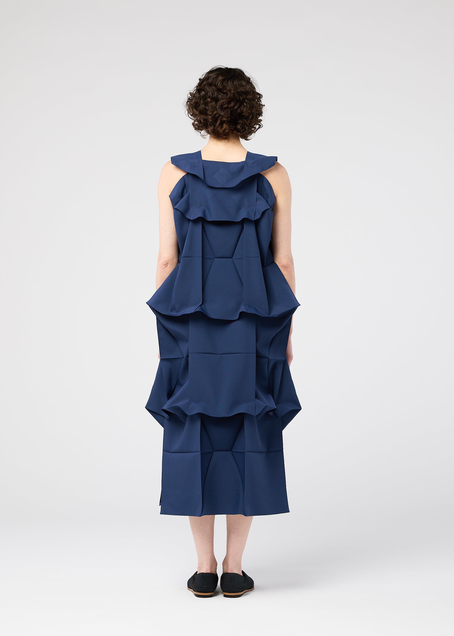 Zoomed view A person with short curly hair faces away wearing the 132 5. ISSEY MIYAKE NO.6 SOLID dress, which features large, structured geometric ruffles and a three-dimensional silhouette, with black slip-on shoes against a white background.