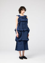 A person with short, curly hair wears the 132 5. ISSEY MIYAKE NO.6 SOLID sleeveless navy dress with geometric layered ruffles, standing against a plain light gray background and paired with black slip-on shoes.