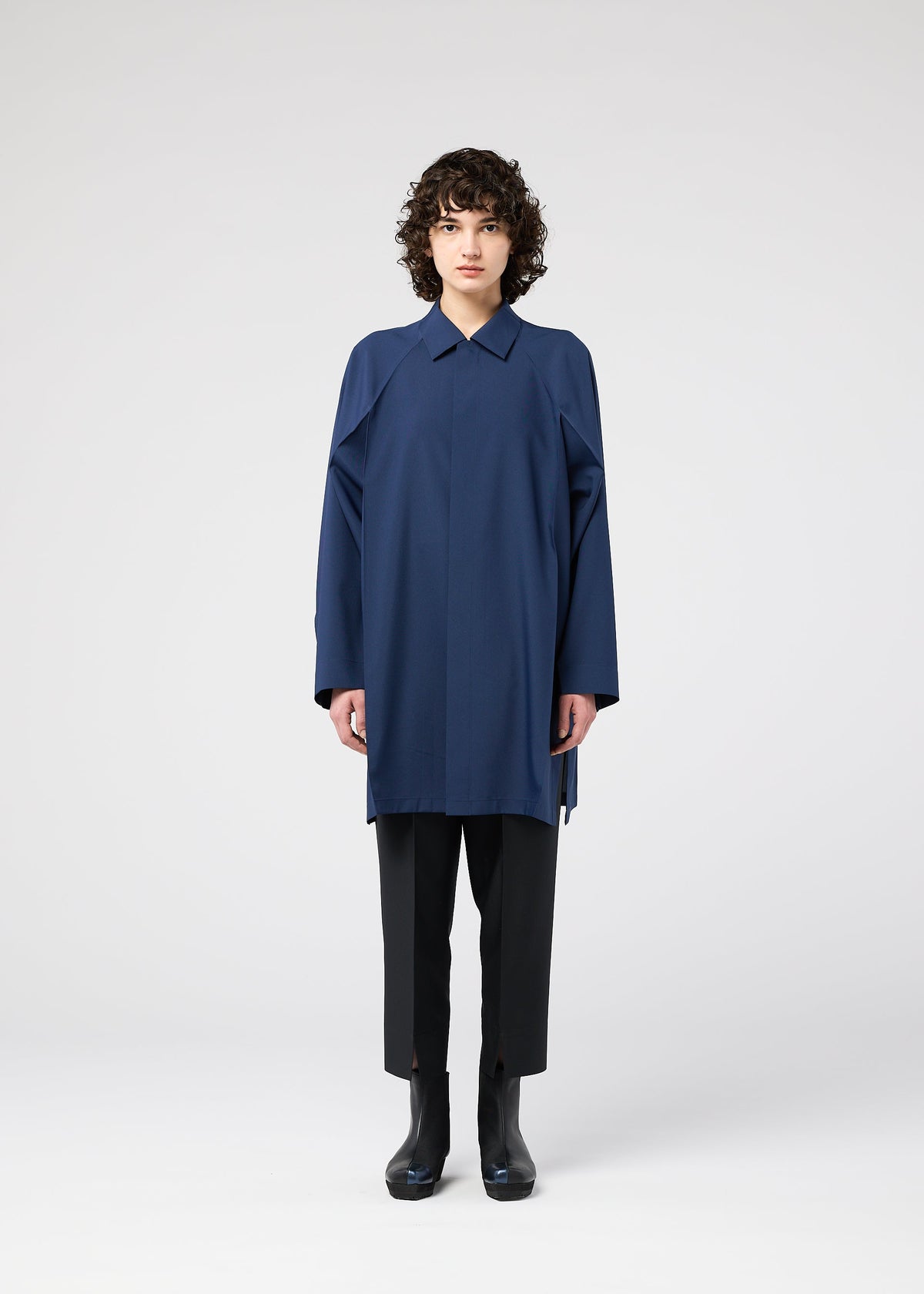 A person with short curly hair faces forward, wearing the FOLD SHIRT by 132 5. ISSEY MIYAKE—a loose blue top with raglan sleeves—paired with black pants and boots, set against a plain white background. view 8 of 12