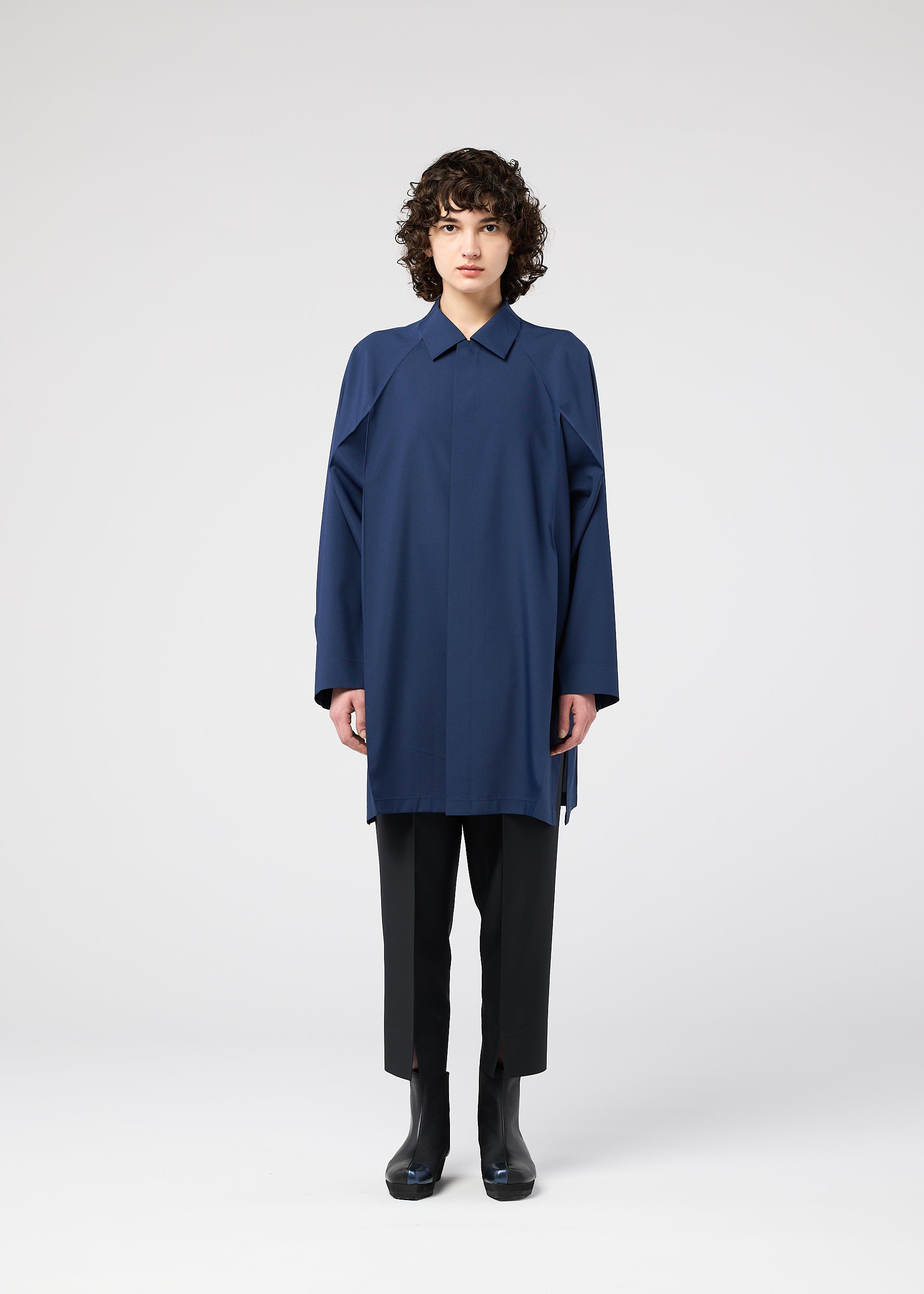 Zoomed view A person with short curly hair faces forward, wearing the FOLD SHIRT by 132 5. ISSEY MIYAKE—a loose blue top with raglan sleeves—paired with black pants and boots, set against a plain white background.