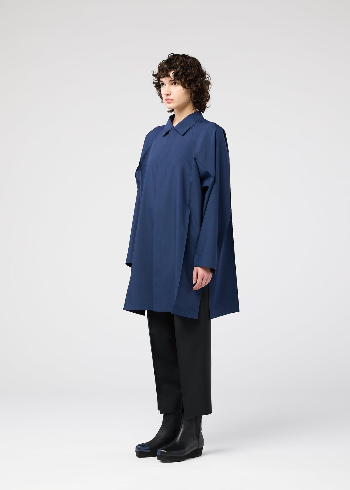 A person with short curly hair stands in profile, wearing the FOLD SHIRT by 132 5. ISSEY MIYAKE, paired with black wide-leg pants and black ankle boots against a plain white background. view 9 of 12