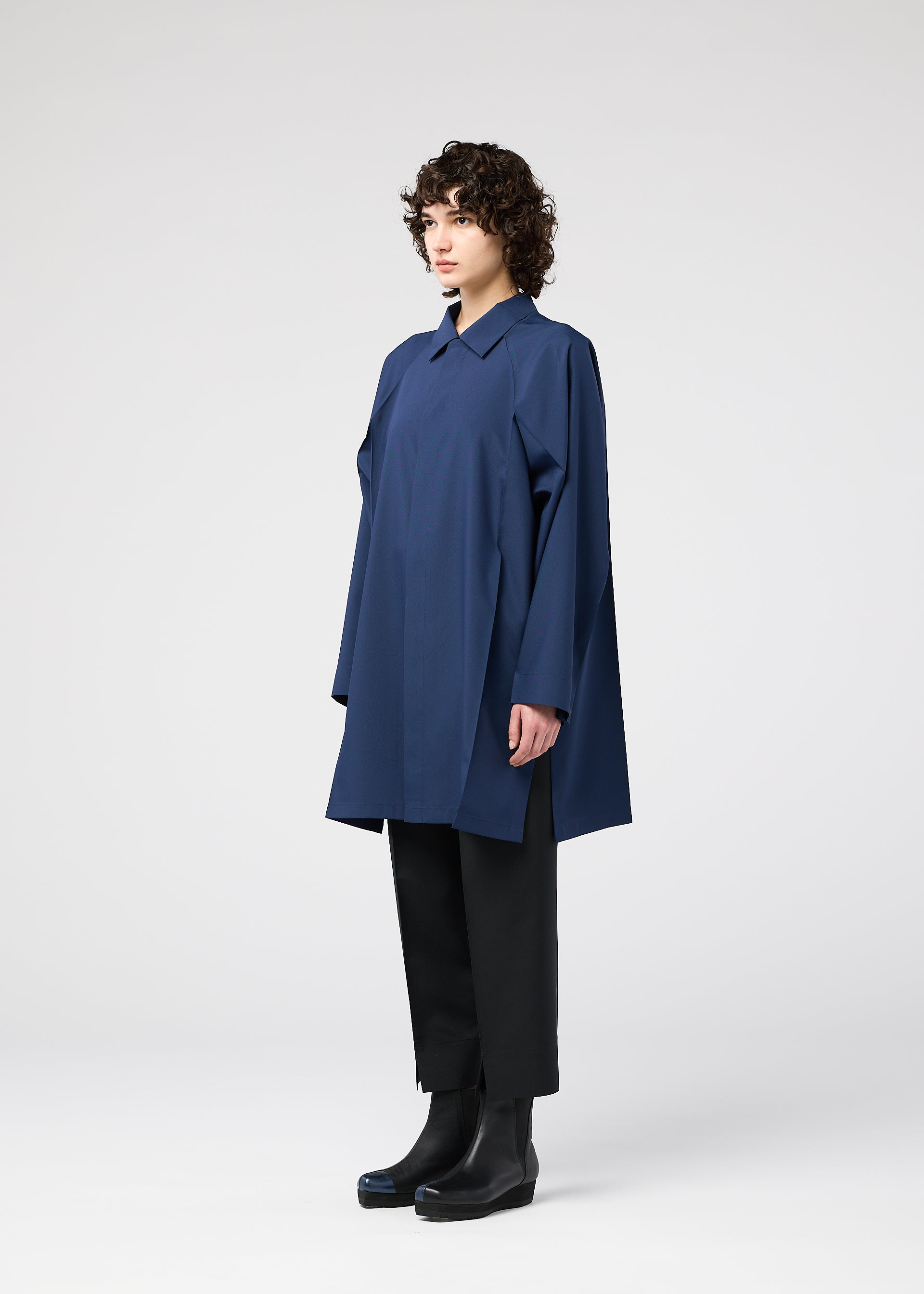 Zoomed view A person with short curly hair stands in profile, wearing the FOLD SHIRT by 132 5. ISSEY MIYAKE, paired with black wide-leg pants and black ankle boots against a plain white background.