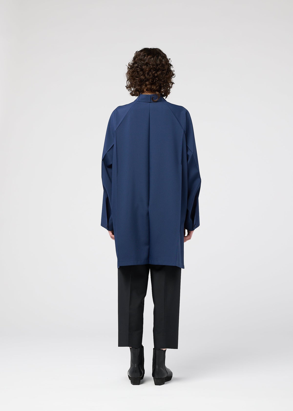 A person with curly hair stands facing away, wearing the FOLD SHIRT by 132 5. ISSEY MIYAKE—a loose-fitting, long blue shirt with raglan sleeves—over black pants and black ankle boots, against a plain white background. view 10 of 12