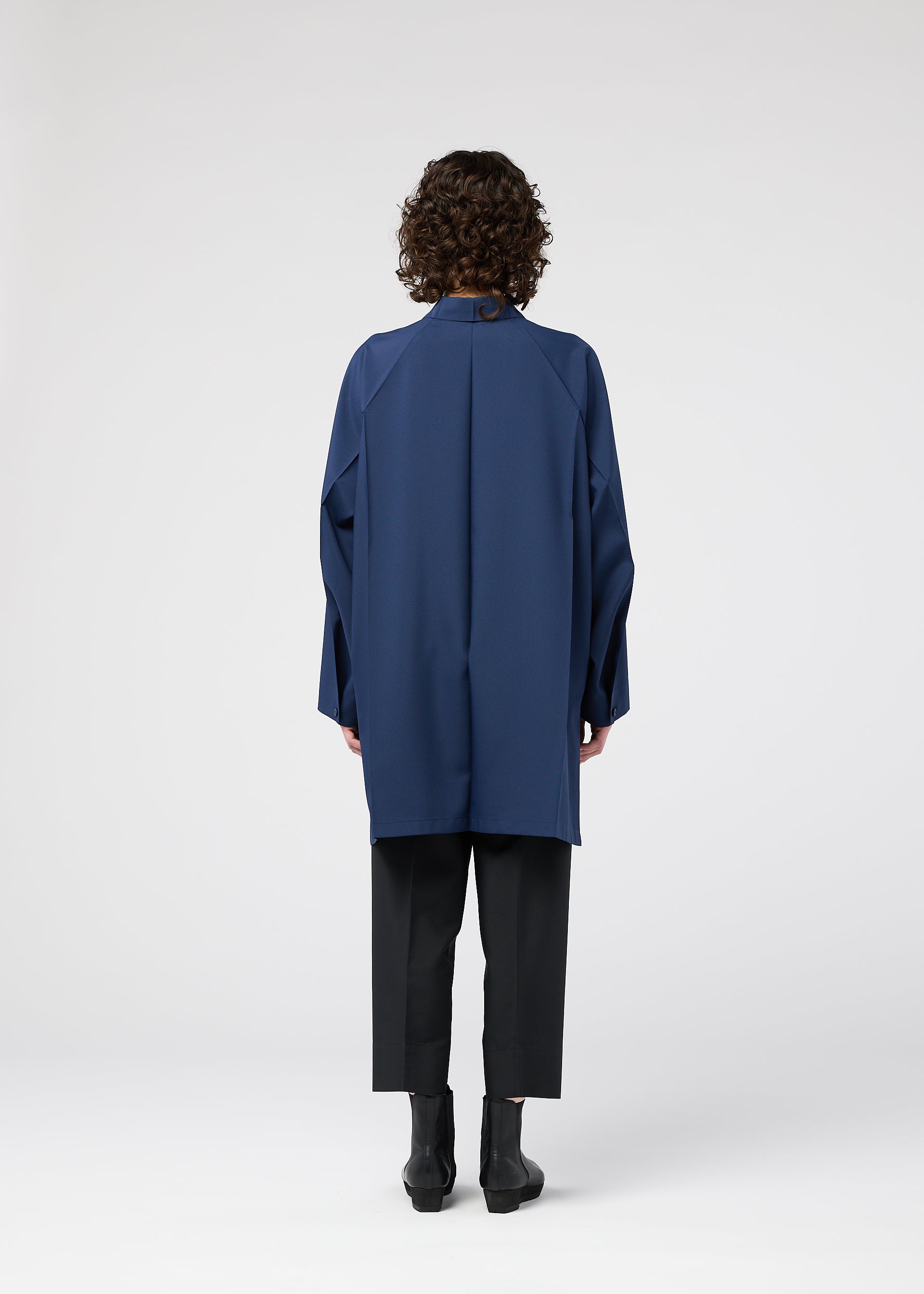 Zoomed view A person with curly hair stands facing away, wearing the FOLD SHIRT by 132 5. ISSEY MIYAKE—a loose-fitting, long blue shirt with raglan sleeves—over black pants and black ankle boots, against a plain white background.