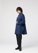 A person with curly hair stands in profile, wearing the oversized navy FOLD SHIRT by 132 5. ISSEY MIYAKE over black pants and black heeled ankle boots, set against a plain light-gray background.